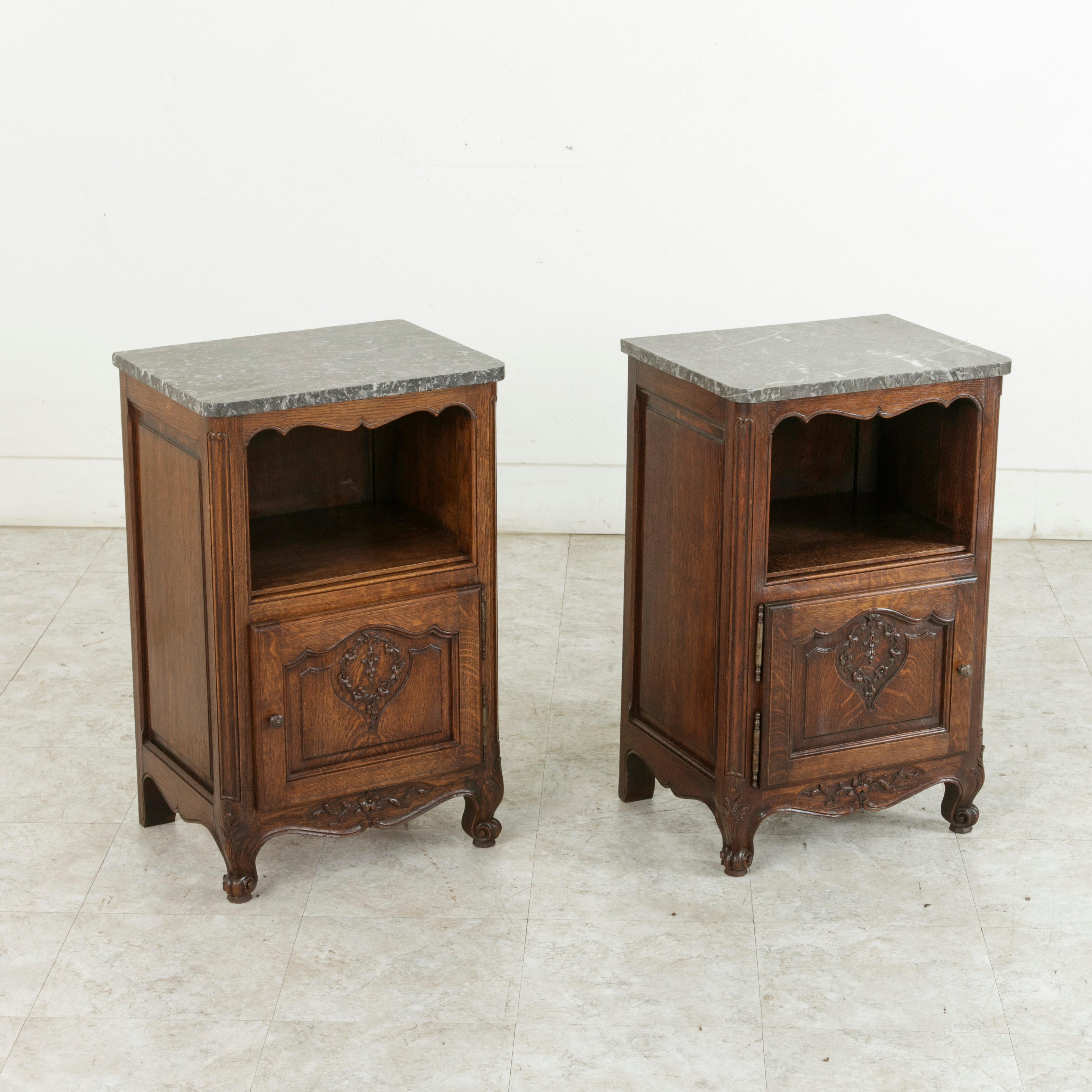 This pair of French Louis XV style oak nightstands from the turn of the 20th century features Saint Anne marble tops with niches below and solid panel sides. The single doors of the lower cabinets are adorned with hand carved wreathes of flowers,