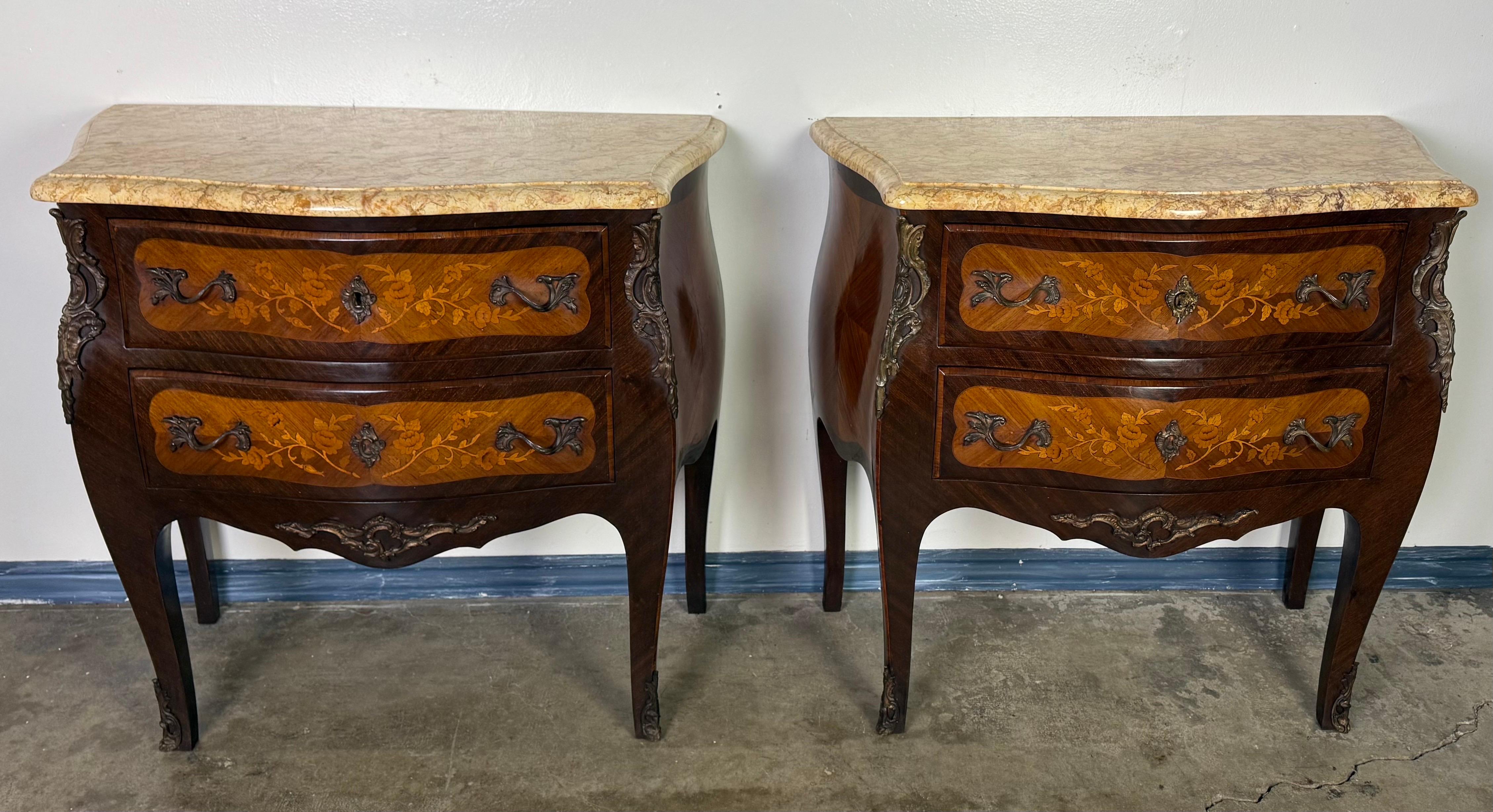 A fine pair of French Louis XV style commodes, each with two drawers, richly inlaid with floral marquetry. The bombe-shaped cases feature veneered surfaces in contrasting woods, highlighted with delicate floral sprays on the drawer fronts. The