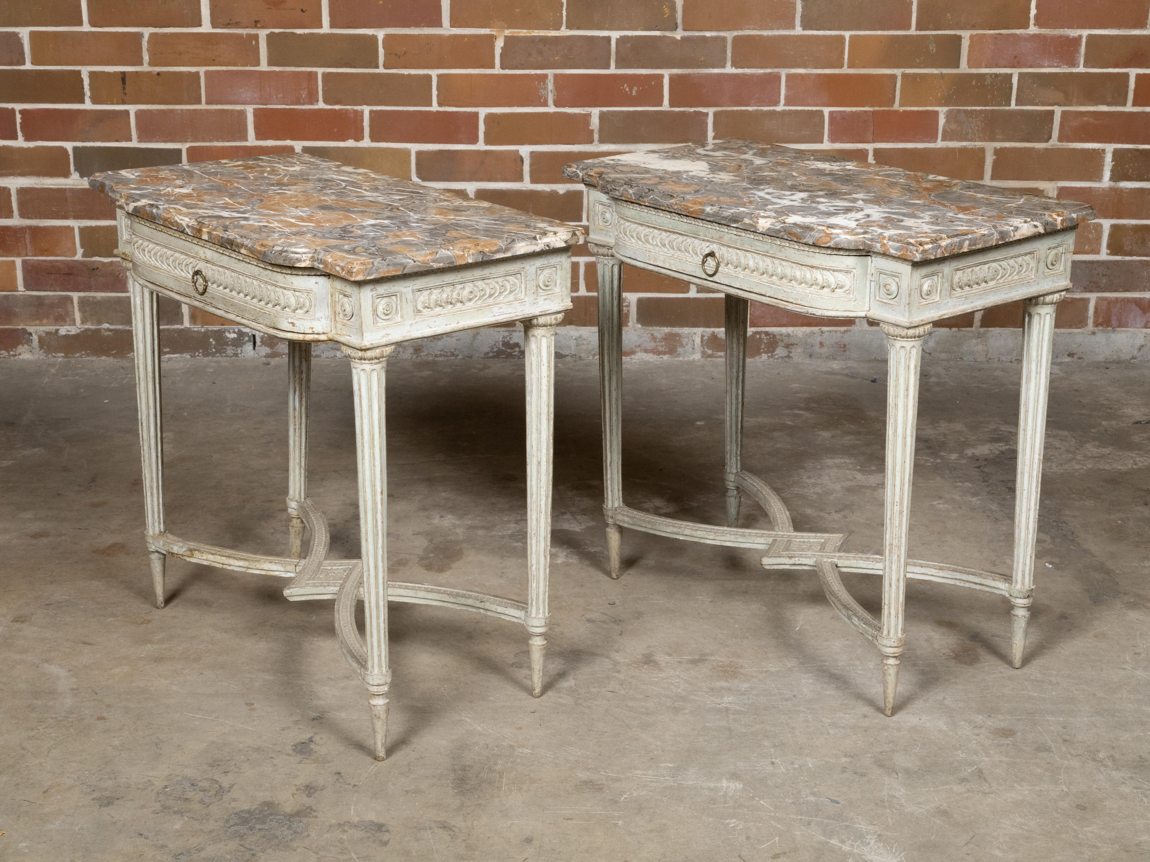 A pair of French Louis XVI period antique white painted console tables from the 18th century with marble tops, carved stacked coin style frieze and fluted legs. This pair of French Louis XVI period console tables, dating back to the 18th century,