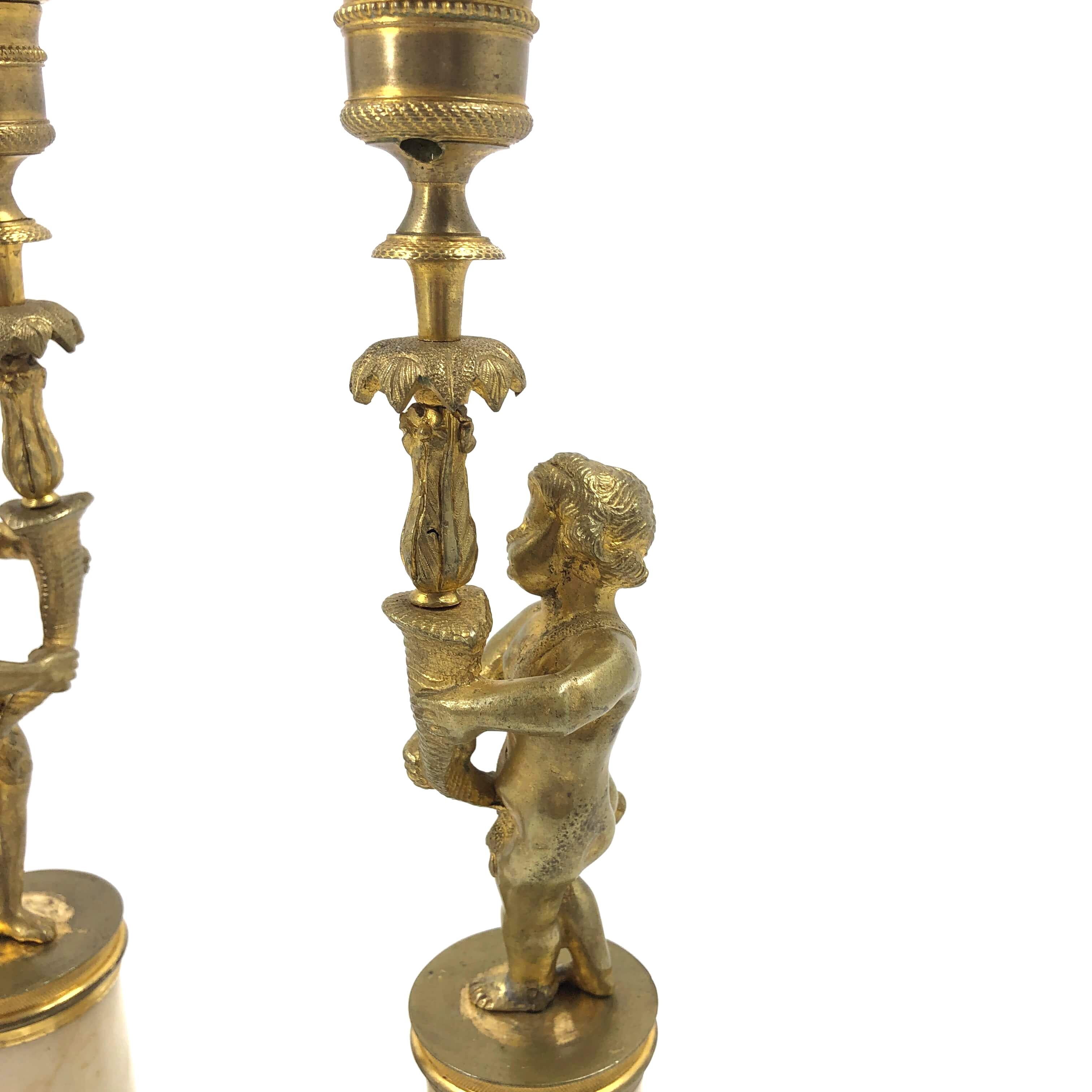 A pair of French Louis XVI figural putti gilt bronze candlesticks. Putti holding cornucopia arms with palm leaves and chased rims, above a neoclassical round marble pedestal with chased bronze capitals and palm leaves, above a square plinth base.