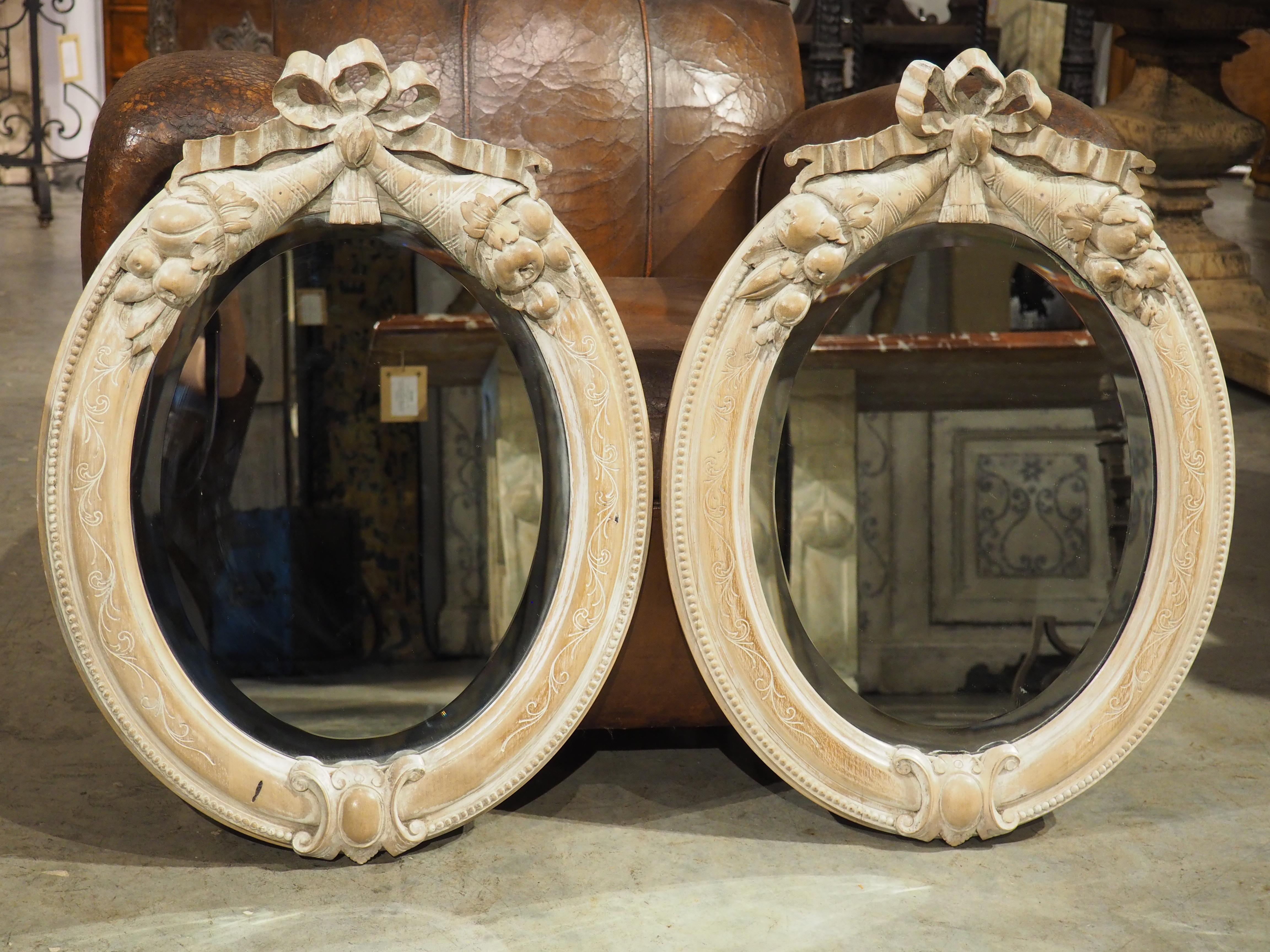 This pair of French wooden mirrors were hand-carved in the style of Louis XVI by an expert-level woodworker in the early 1900s. Perhaps the most eye-catching motif is the crest of each frame, which is comprised of a pair of overladen cornucopias