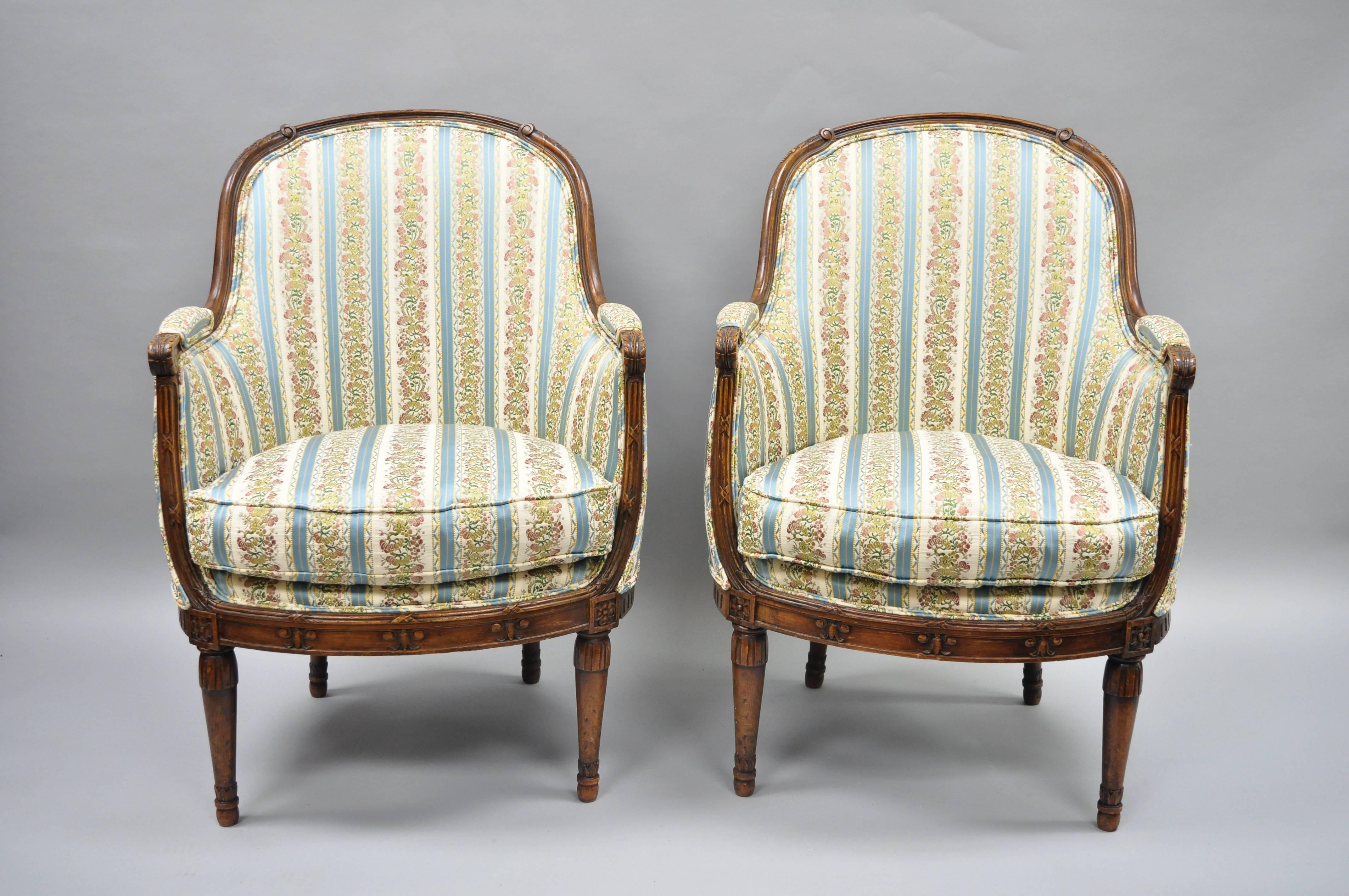 Pair of French Louis XVI Directoire style walnut bergere armchairs. Item features finely carved walnut frames, bell flower and drape details, reeded legs, blue, cream and pink silk upholstery, partial down fill cushions, upholstered armrests, and