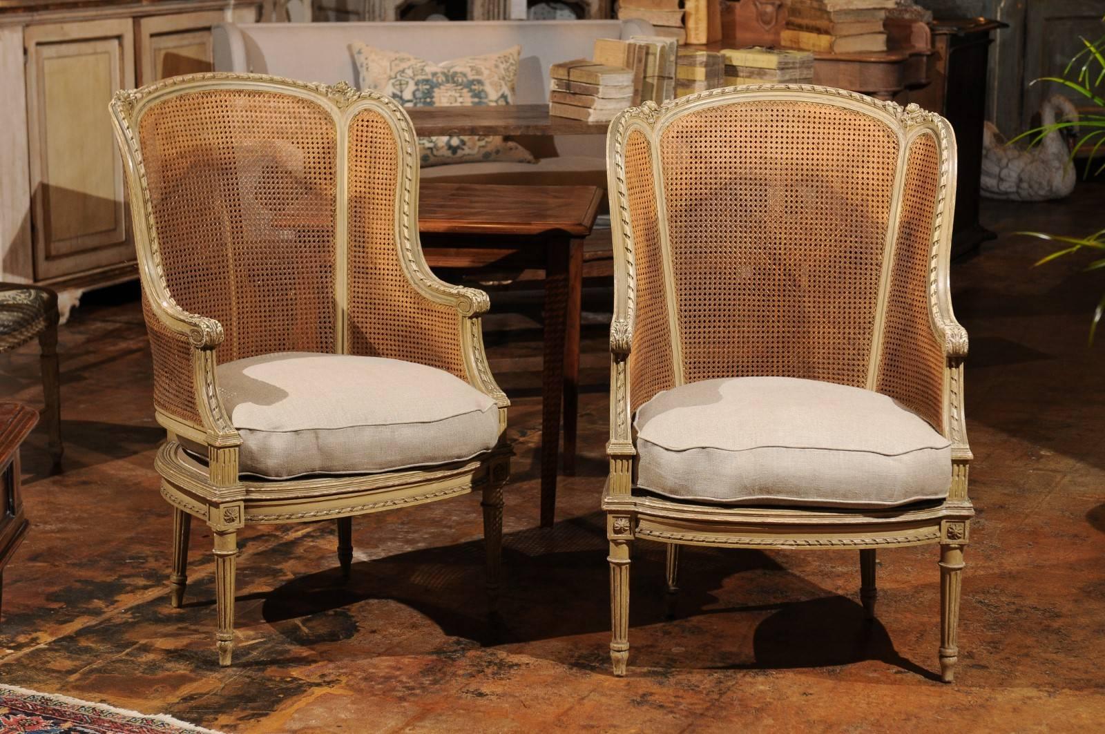 A pair of French Louis XVI style painted wood and cane bergères chairs from the late 19th century. Each of this pair of bergères à oreilles features a caned winged back, adorned on the upper rail with a twisted ribbon motif, accented with delicate