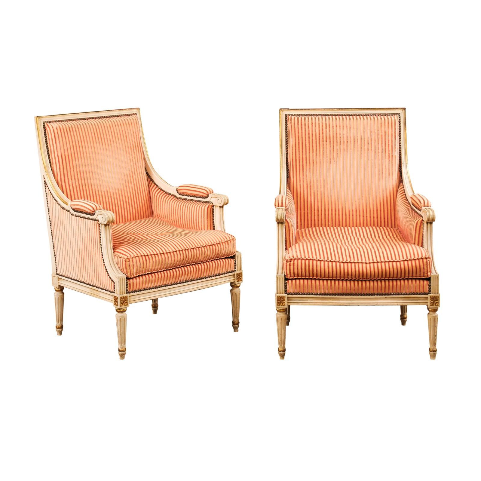 A pair of French Louis XVI style painted wood bergères à la Reine chairs from the early 20th century, with carved rosettes, gilded details and old upholstery. Created in France at the Turn of the Century which saw the transition between the 19th to
