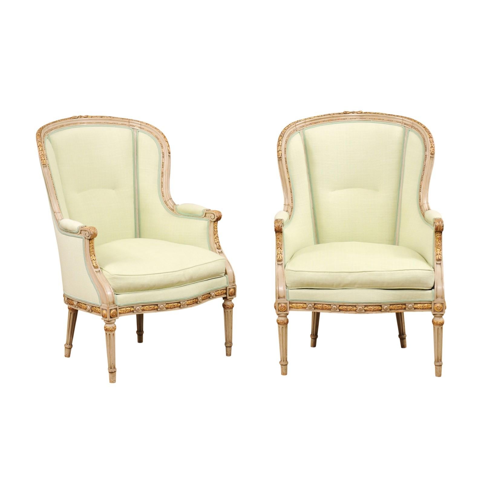 A pair of French Louis XVI style painted wood, parcel-gilt bergères chairs from the early 20th century with soft yellow green double welt upholstery, scrolled arms, carved foliage and fluted legs. Created in France at the Turn of the Century which