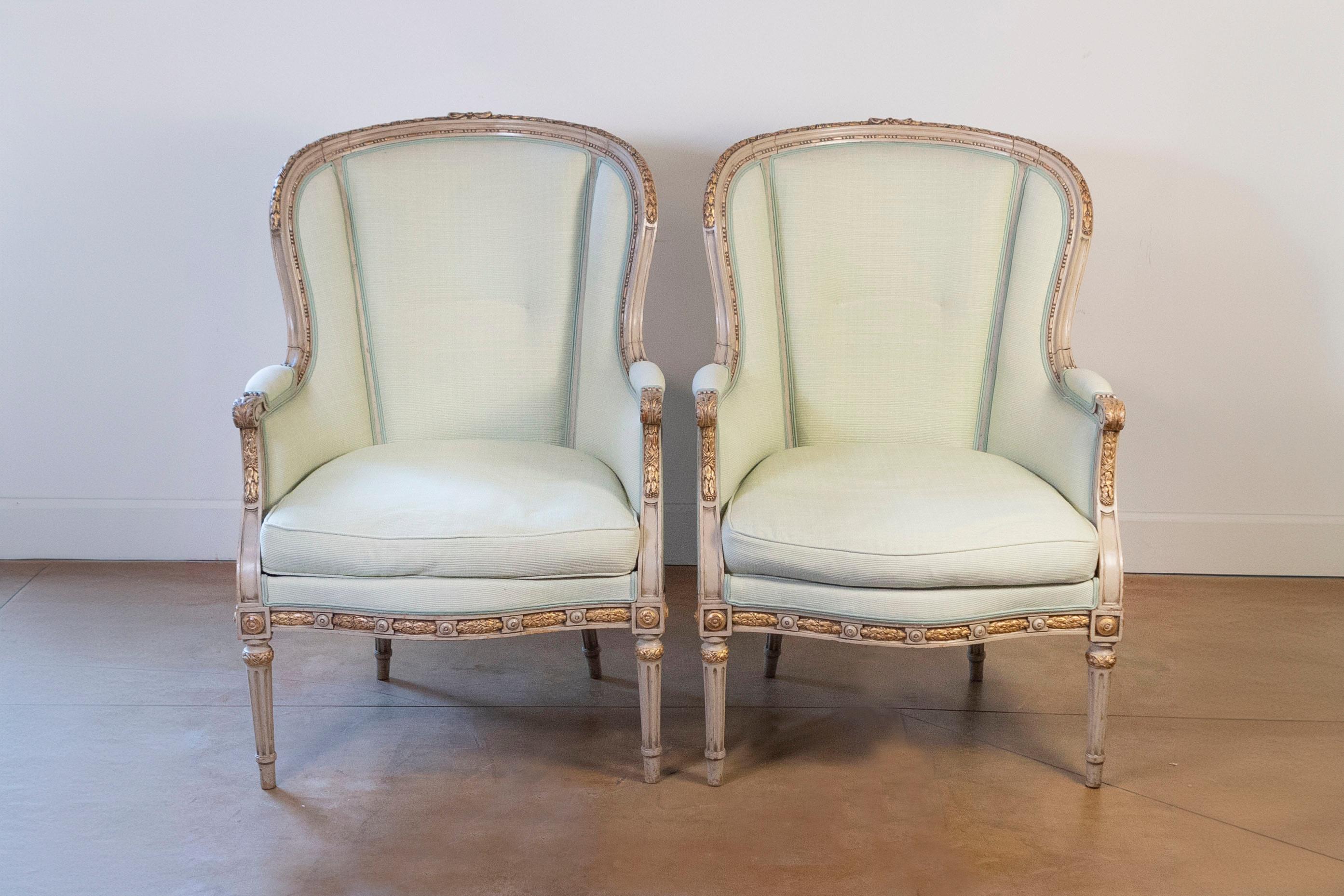A pair of French Louis XVI style painted wood, parcel-gilt bergères chairs from the early 20th century with soft yellow green double welt upholstery, scrolled arms, carved foliage and fluted legs. Created in France at the Turn of the Century which