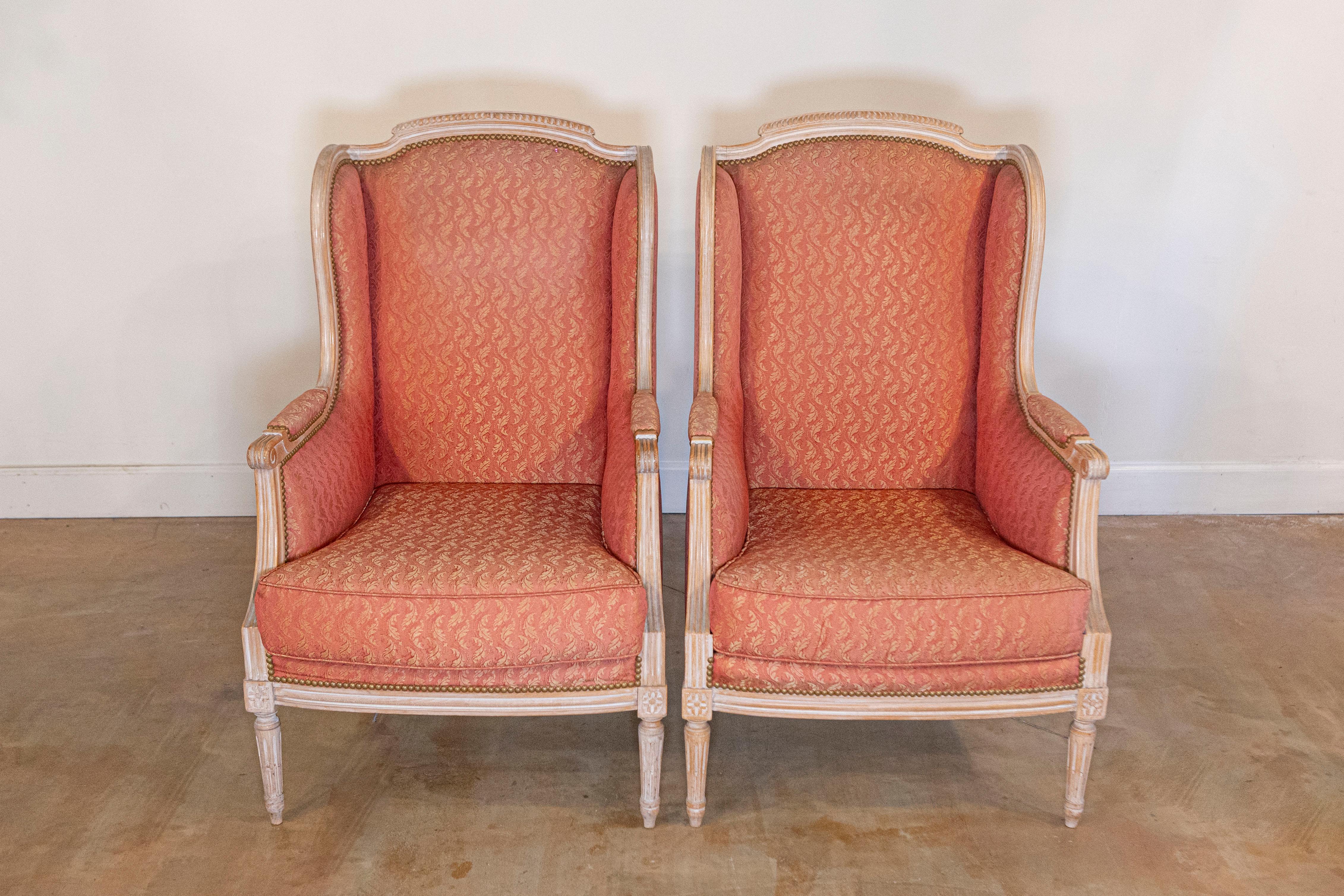 A pair of French Louis XVI style painted wood bergère chairs from the early 20th century, with carved décor and foliage-themed upholstery. Created in France at the Turn of the Century, this pair of bergères showcases the stylistic characteristics of