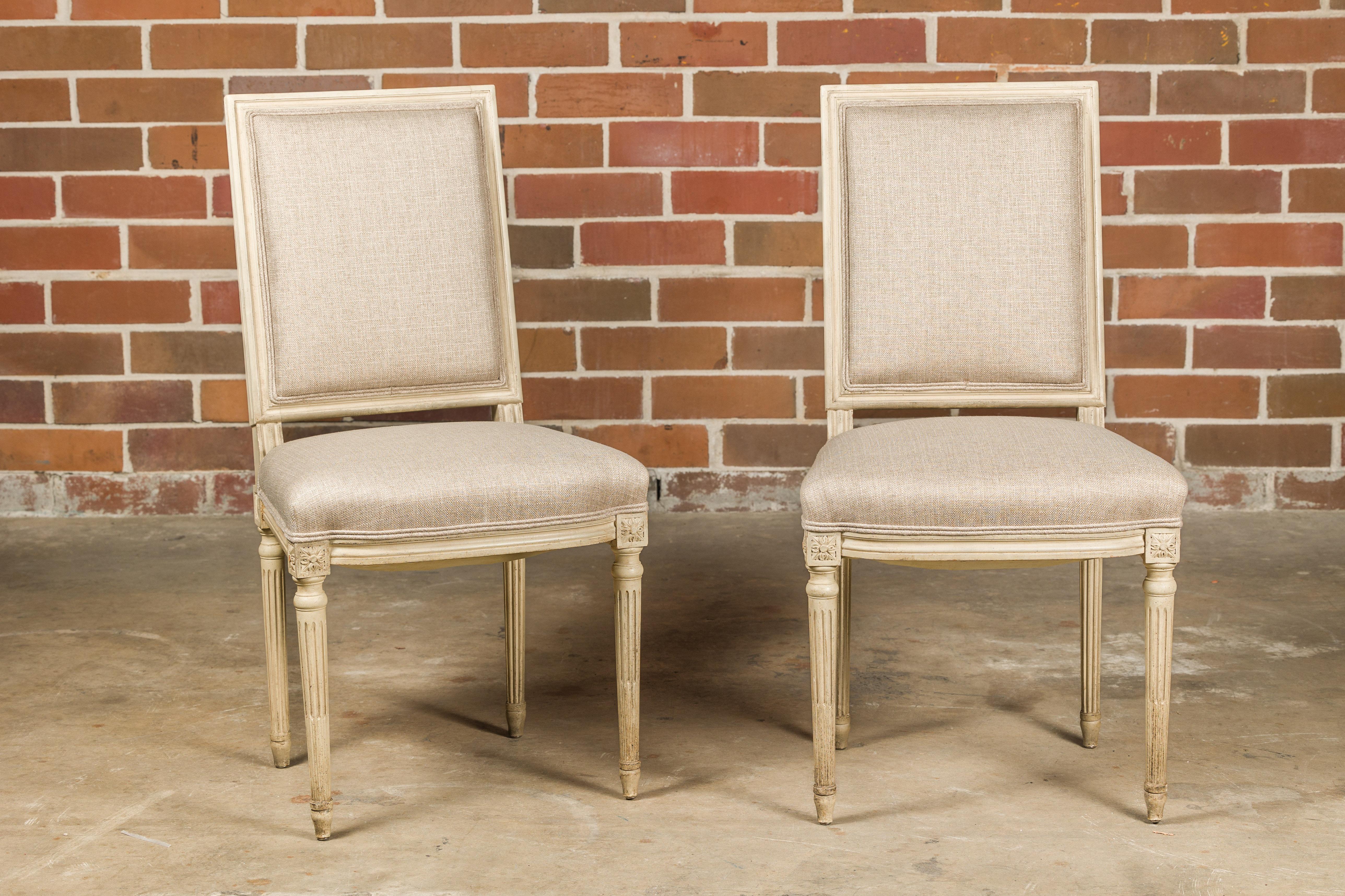 A pair of French Louis XVI style painted side chairs from circa 1920 with rectangular backs, fluted legs, carved rosettes and linen upholstery. This exquisite pair of French Louis XVI style painted side chairs from circa 1920 exudes timeless