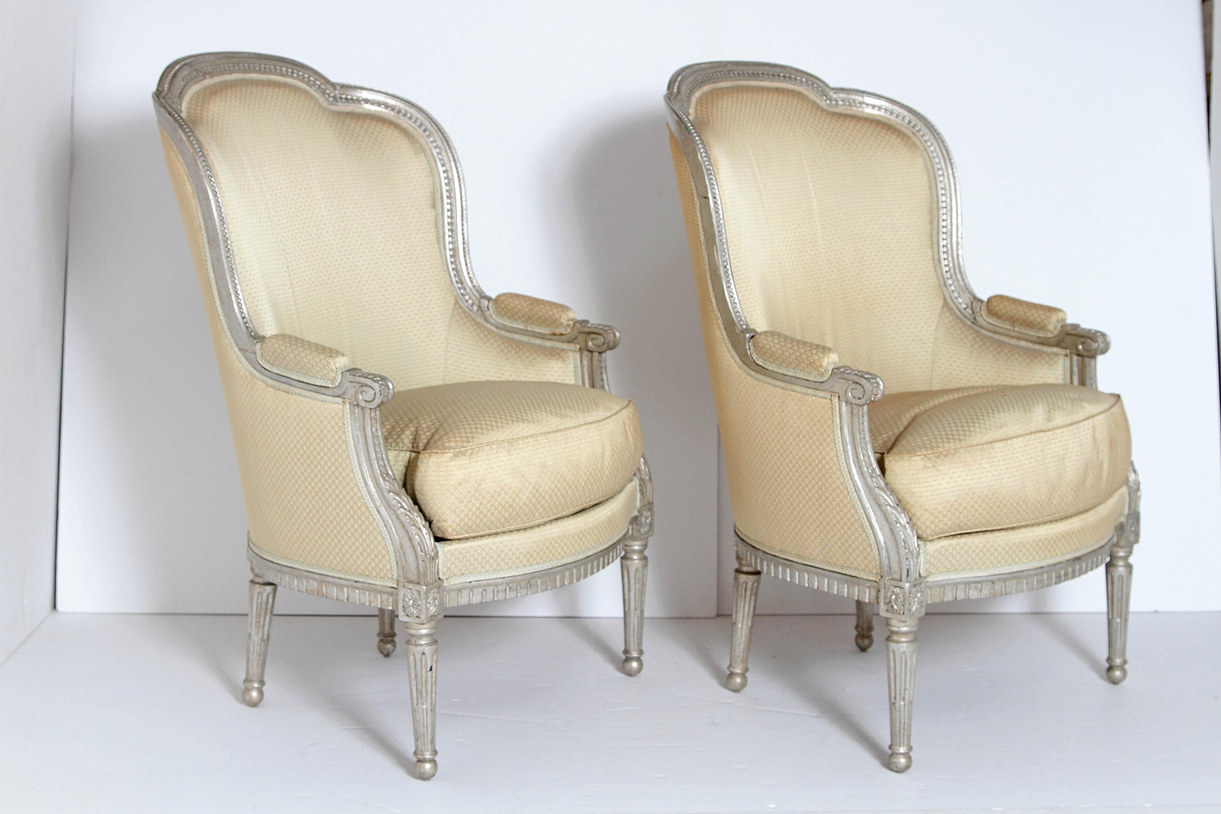 A pair of French Louis XVI style bergères with a nicely carved silver gilt frames. Loose upholstered seat cushions and padded arm rests, round fluted tapered legs, late 19th century, France.