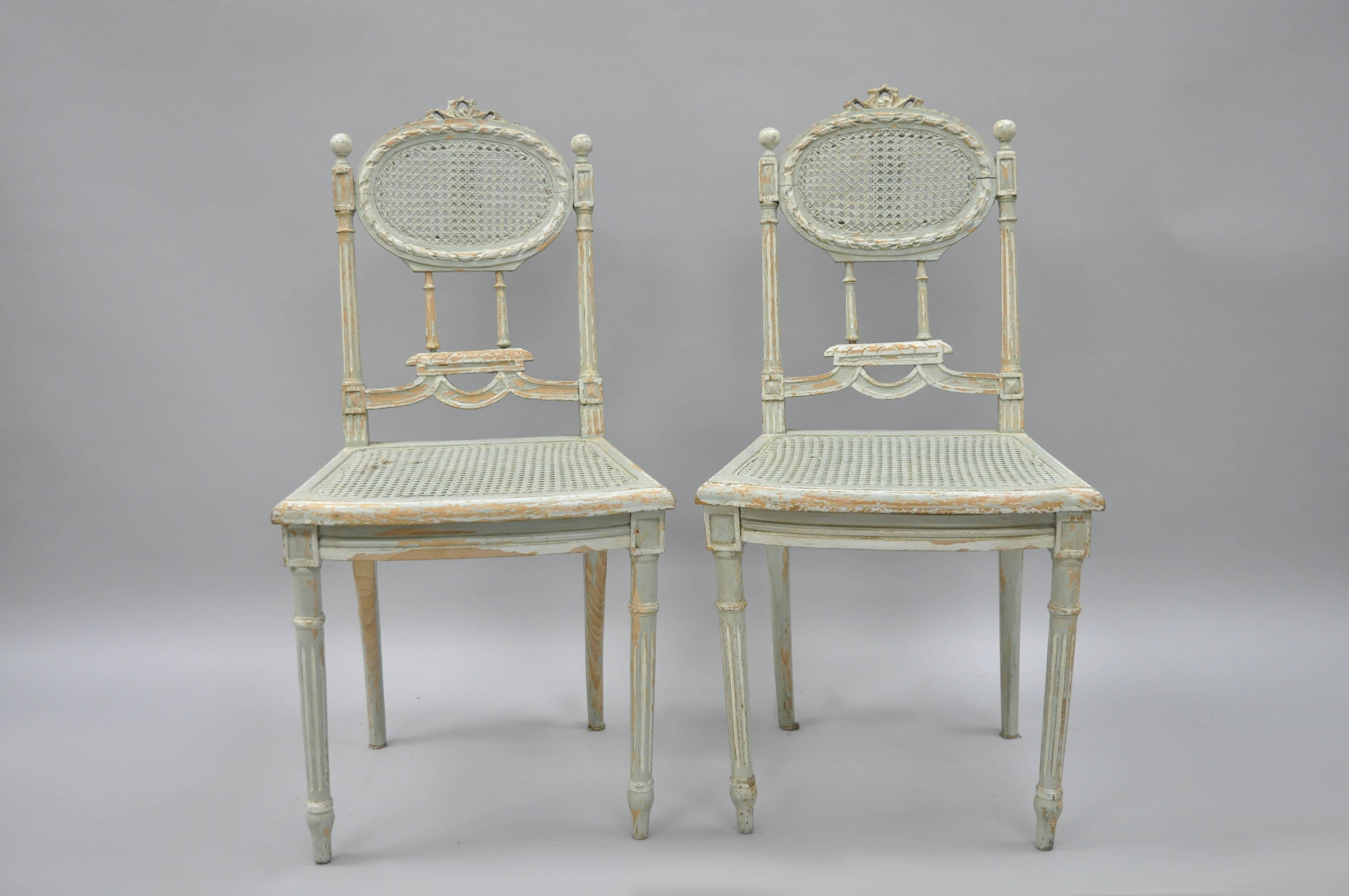 Pair of distress painted French Louis XVI style side chairs. Chairs are constructed of solid wood with reeded and tapered legs, carved bow tie crest and other classic French carvings throughout. Backs and seats are caned and the original painted