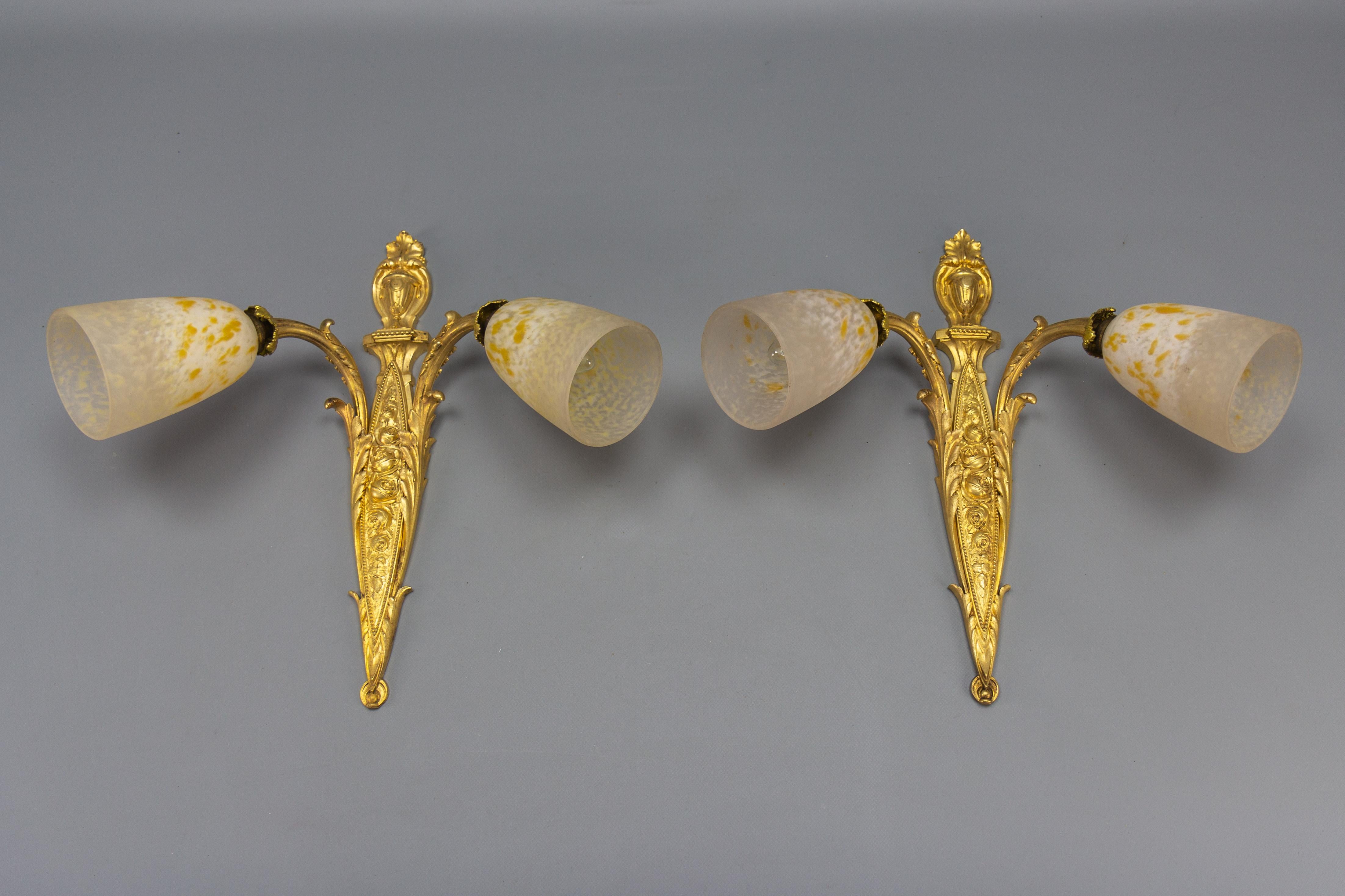 Pair of French Louis XV style bronze and white and yellow glass twin arm sconces from ca. 1910.
These impressive Louis XVI-style sconces feature bronze arms and a backplate of characteristic Louis XVI or Neoclassical-style design, richly decorated