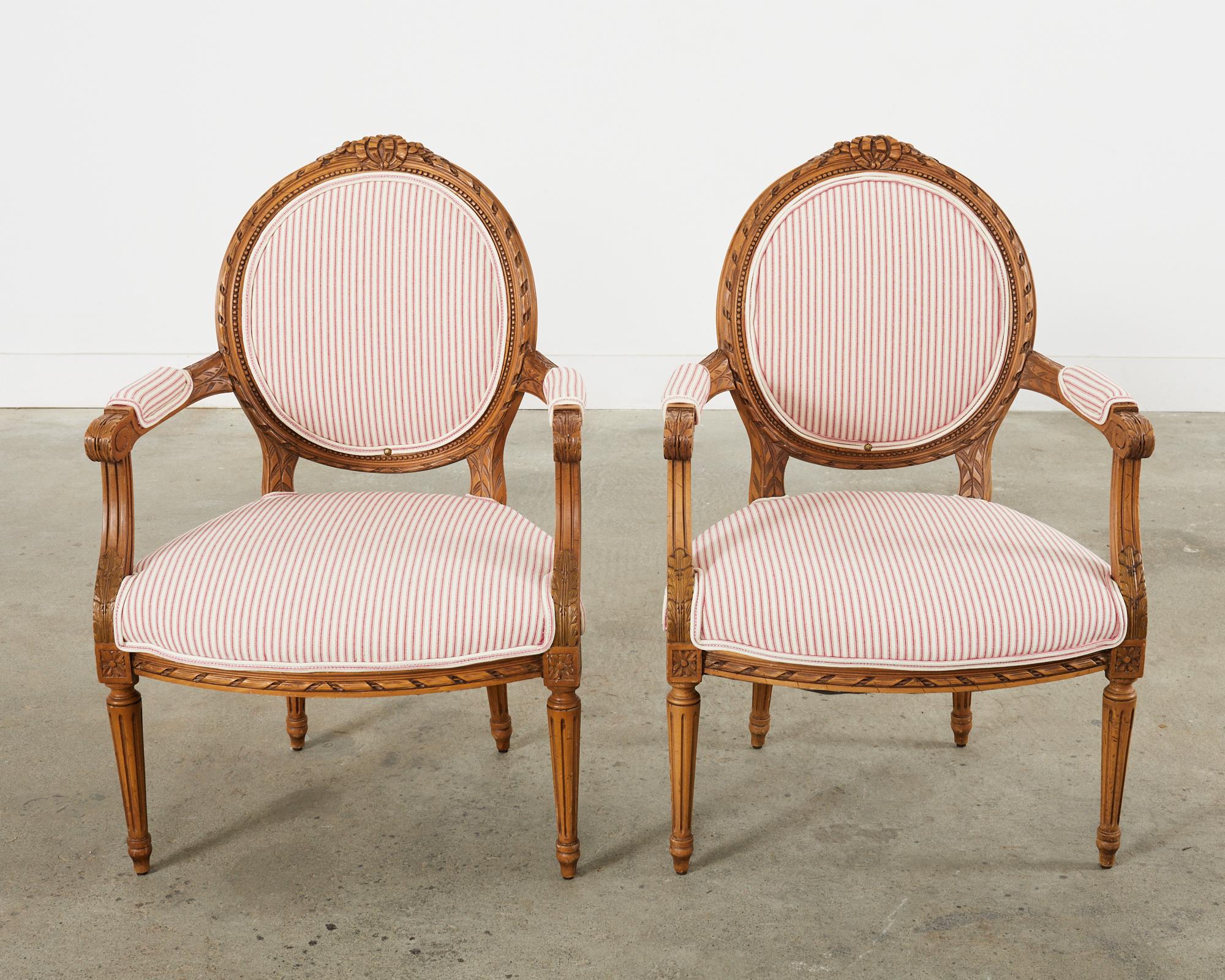 Gorgeous pair of French carved fauteuil armchairs made in the grand Louis XVI taste. The intricately carved frames are decorated with neoclassical motifs of acanthus, rosettes, and ribbon swag bows. The chairs have wide arms measuring 25 inches high