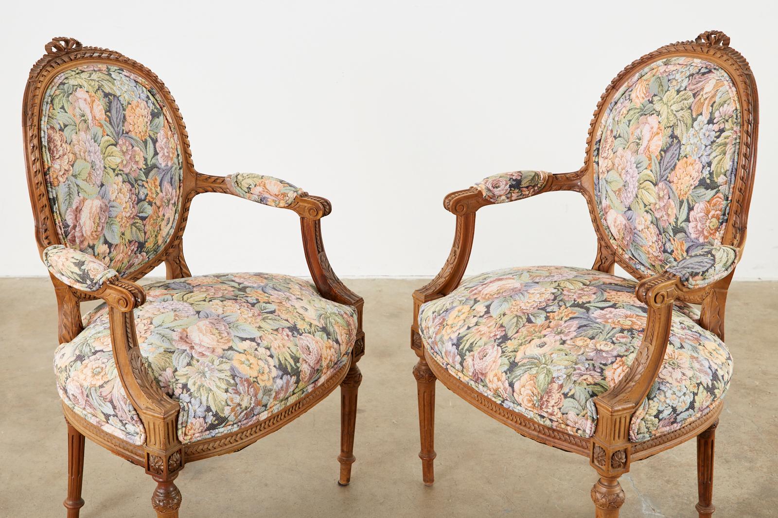 Impressive pair of French carved fauteuil armchairs made in the Louis XVI taste. The chairs feature elaborately hand carved frames with rope designs, rosettes, acanthus, and ribbon bow crests on top. Upholstered with a tapestry style fabric with