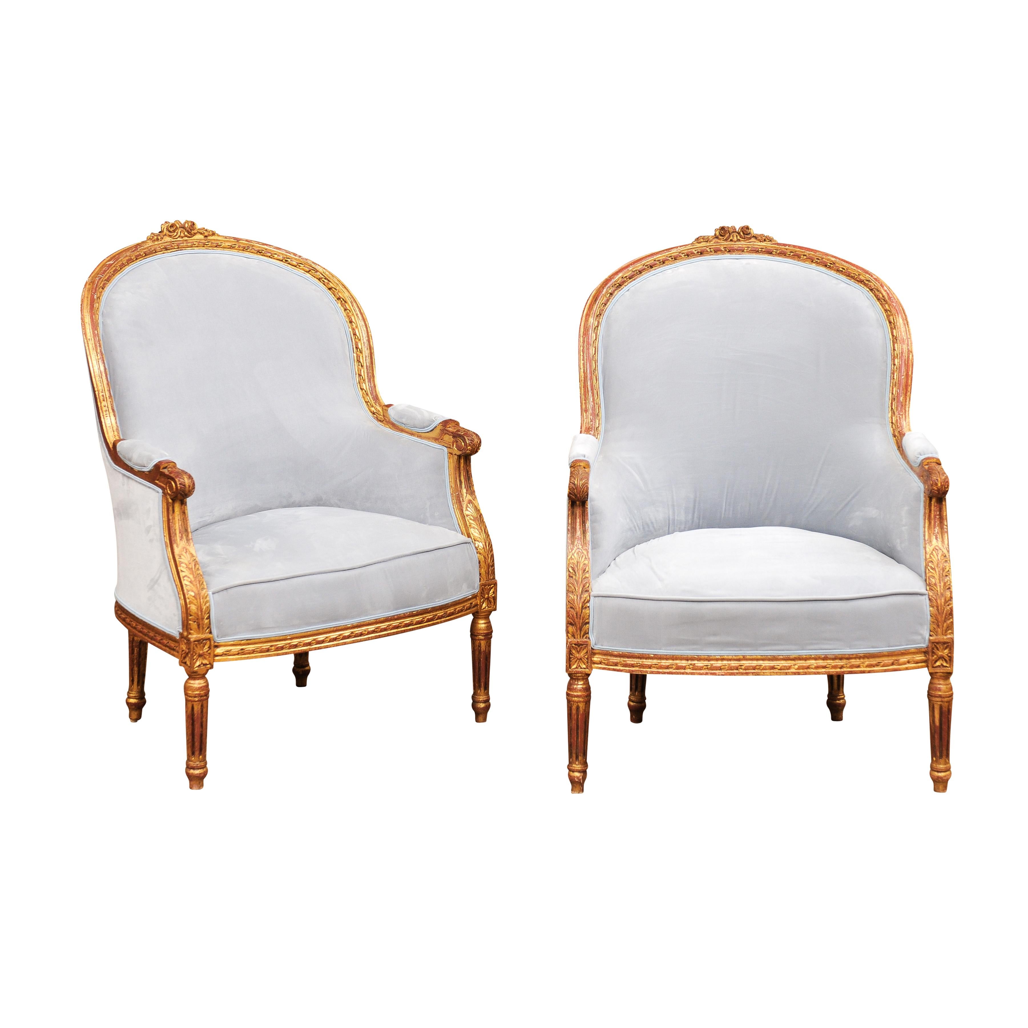 A pair of French Louis XVI style bergères chairs from the 20th century, upholstered with sky blue velvet Pierre Frey fabric with curving backs, floral carved crests, scrolling knuckles, carved acanthus leaves and fluted legs. Indulge in the