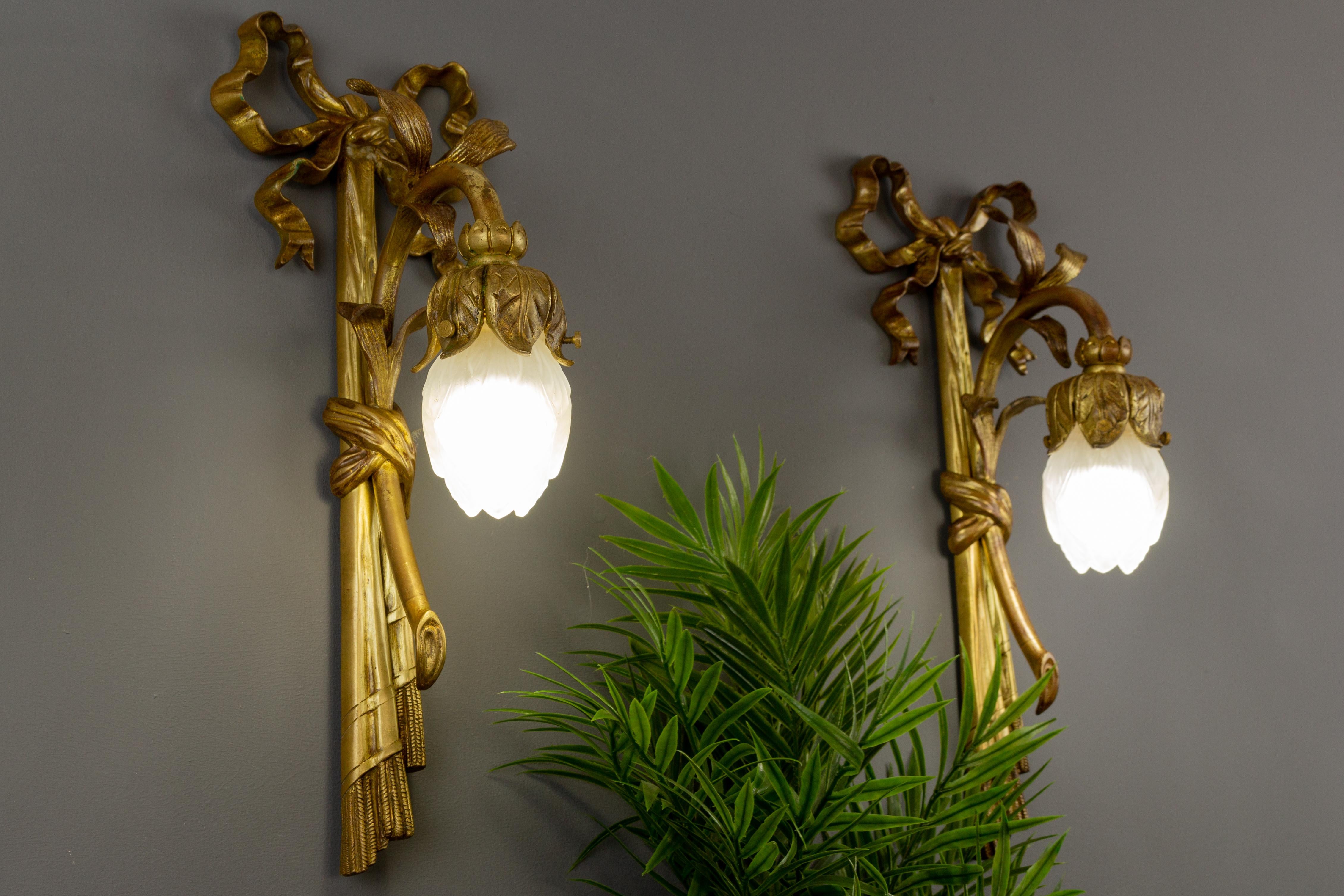 An adorable pair of French Louis XVI-style gilded bronze wall lights in the shape of a flower, tied with a beautiful ribbon and tassel on top of each sconce.
Each has a white frosted glass lampshade in a shape of a flower bud and one socket for an