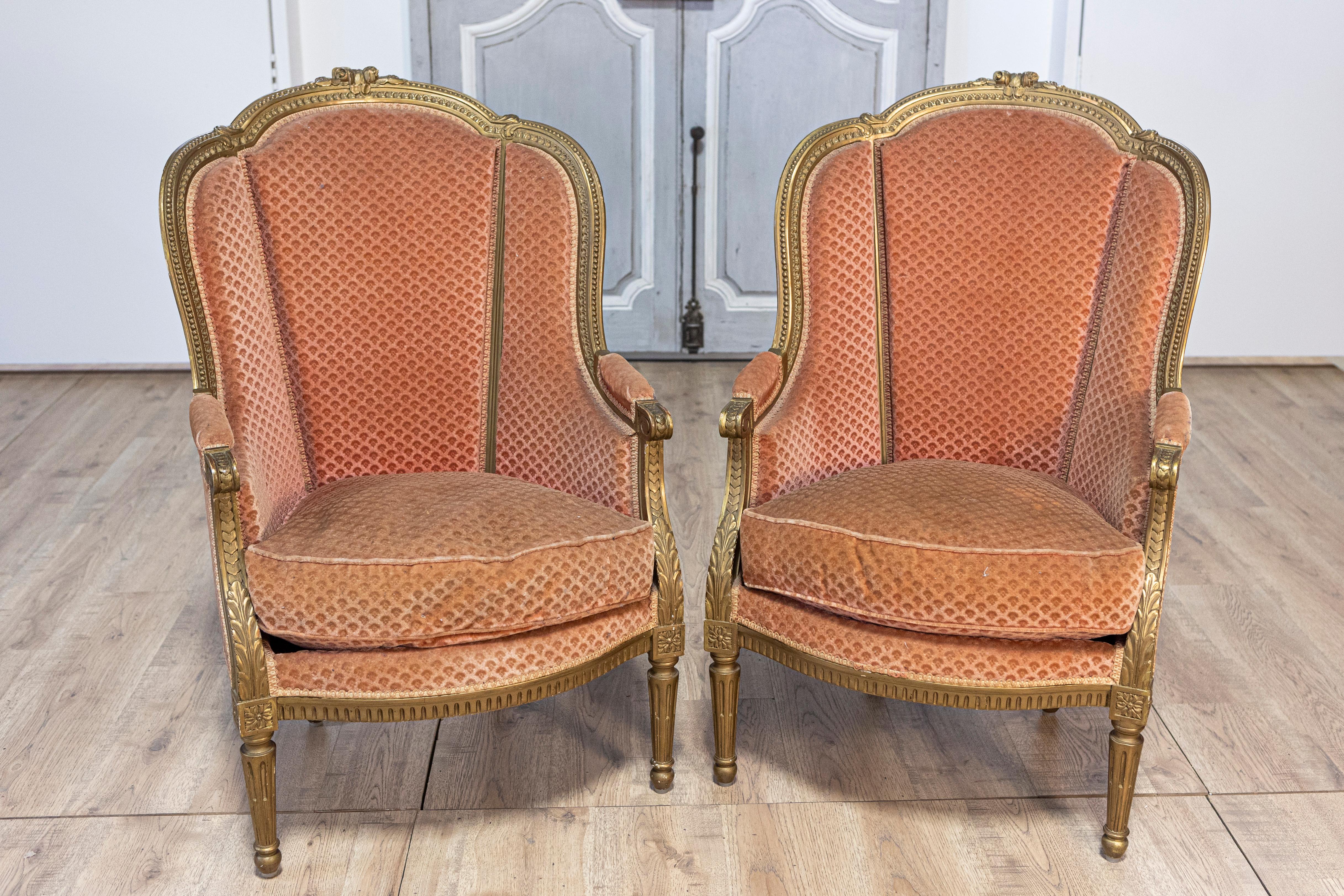 A pair of French Louis XVI style gilded wood bergères chairs from the 20th century with carved décor and fluted legs. Crafted in France during the 20th century, this exquisite pair of French Louis XVI style gilded wood bergère chairs showcases a