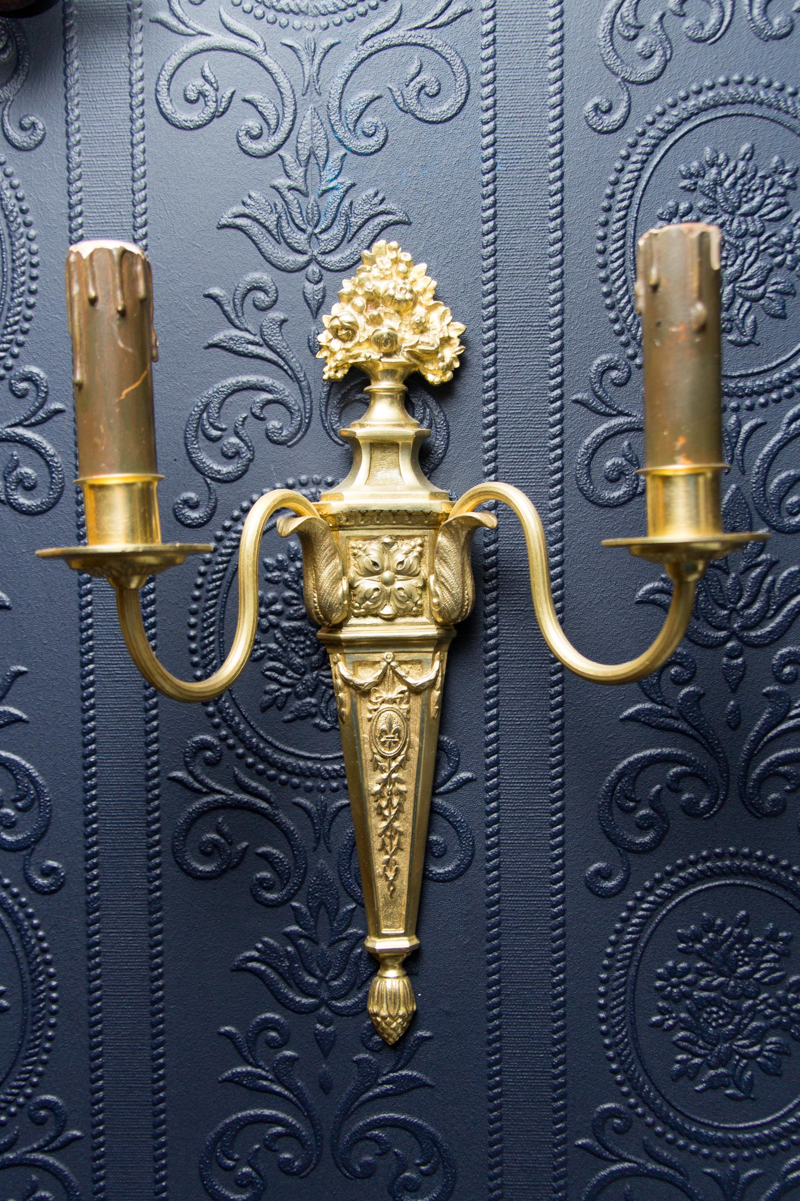 A pair of French early 20th century Louis XVI or neoclassical style gilt bronze sconces. Wall lights have been electrified. Richly decorated with bronze details, like flowers, acanthus leaves, royal French lily, fleur-de-lis, symbol.
Each sconce has