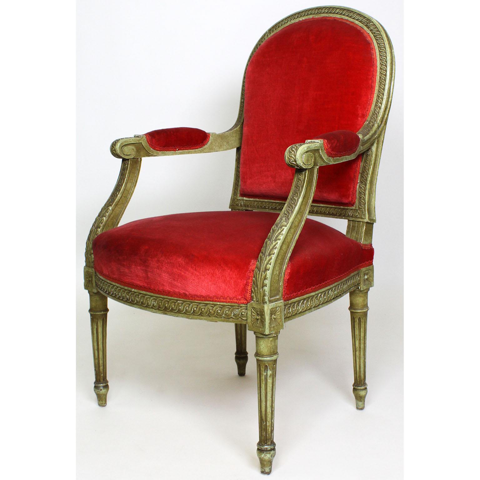 A pair of French Louis XVI Style giltwood carved lacquered Fauteuils (armchairs). The elegantly carved wood frames with rounded backrests, open and padded armrests and fluted legs, in a white/green craquelure finish and upholstered in red velvet.