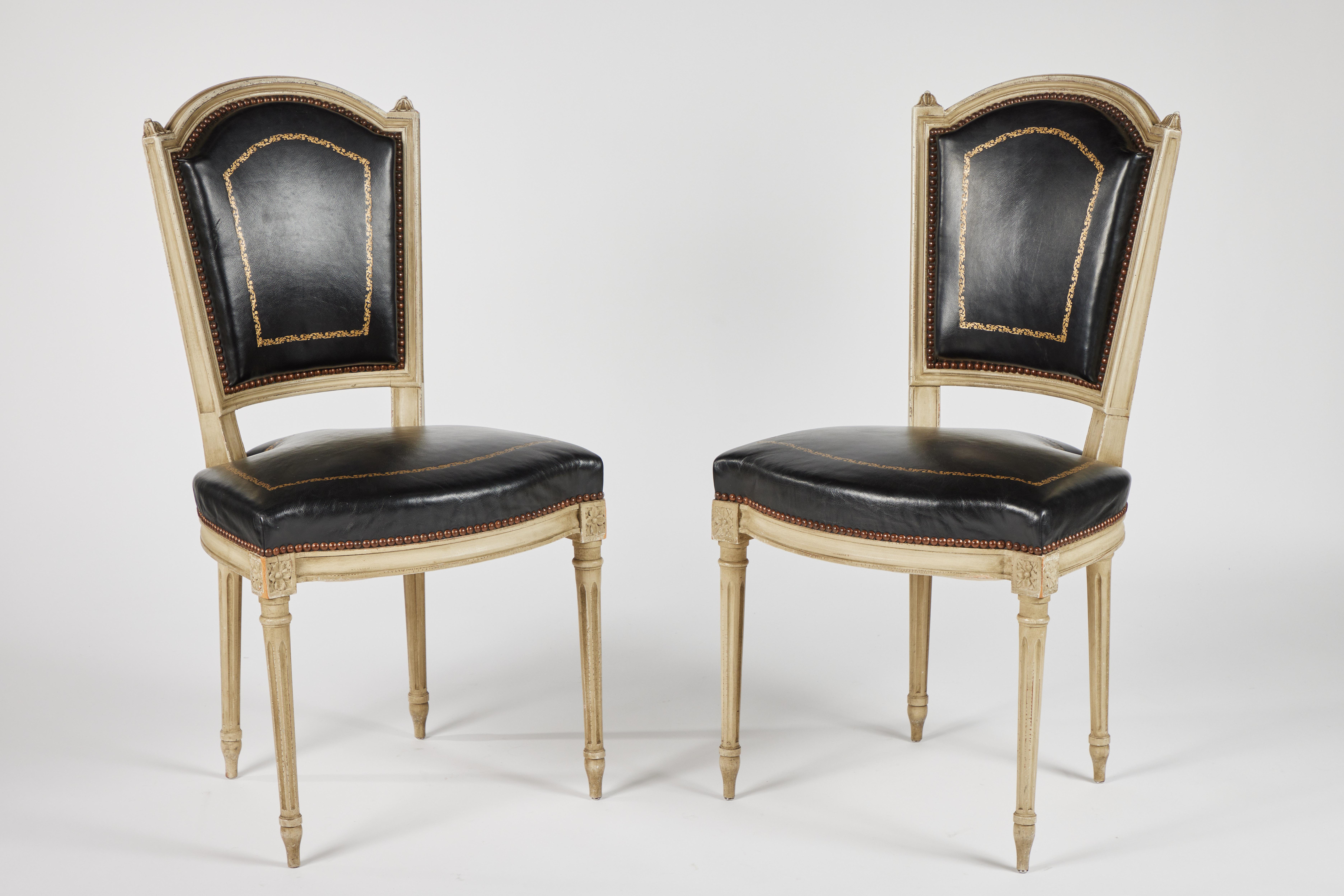 A pair of French Louis XVI style carved and painted dining chairs circa 1940. Black leather upholstered backs and seats supported on turned and fluted legs; gold leaf foliate borders on both seats and backs. The back rest features a dome top flanked