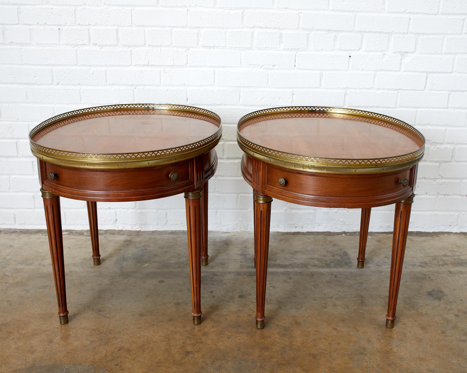 Elegant matching pair of oval Bouillotte side tables or drinks tables made in the French Louis XVI taste. Features a pierced brass galleried edge and brass mounts on the tapered legs ending with brass sabot feet. Each table has a drawer on one end