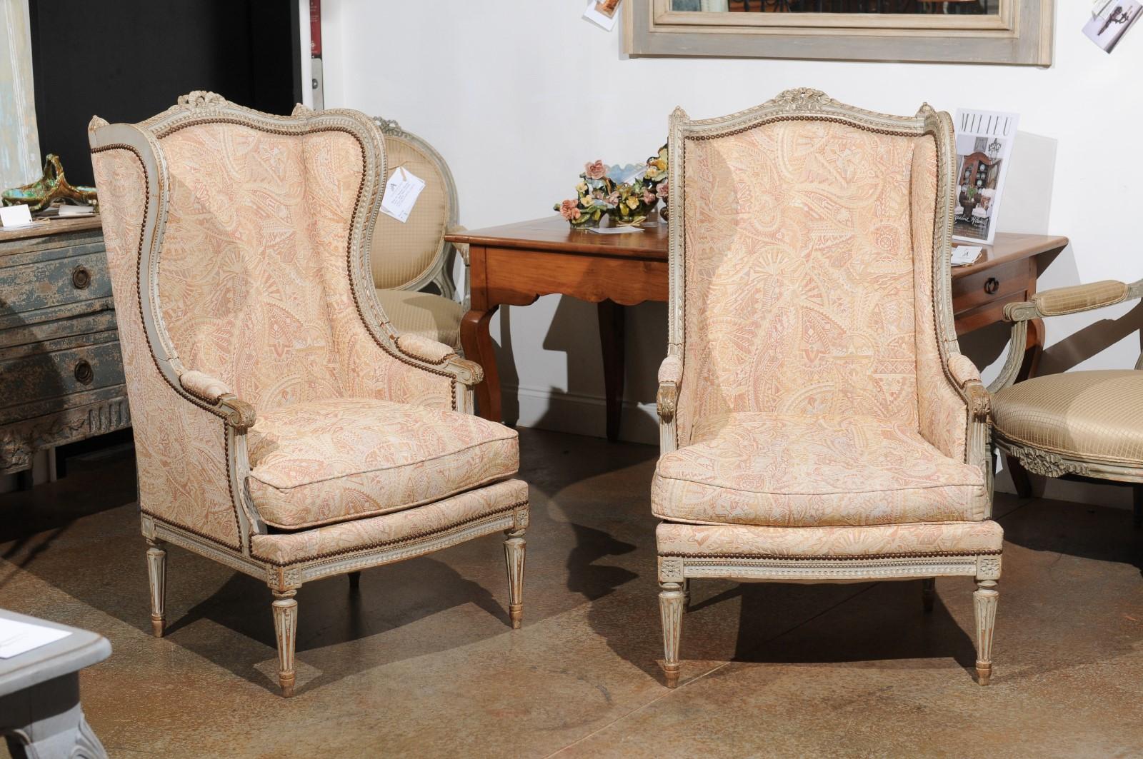 A pair of French Louis XVI style painted wood bergères à oreilles from the 19th century, with carved ribbon motifs and upholstery. Created in France during the 19th century, each of this pair of bergères features a wingback topped with a carved