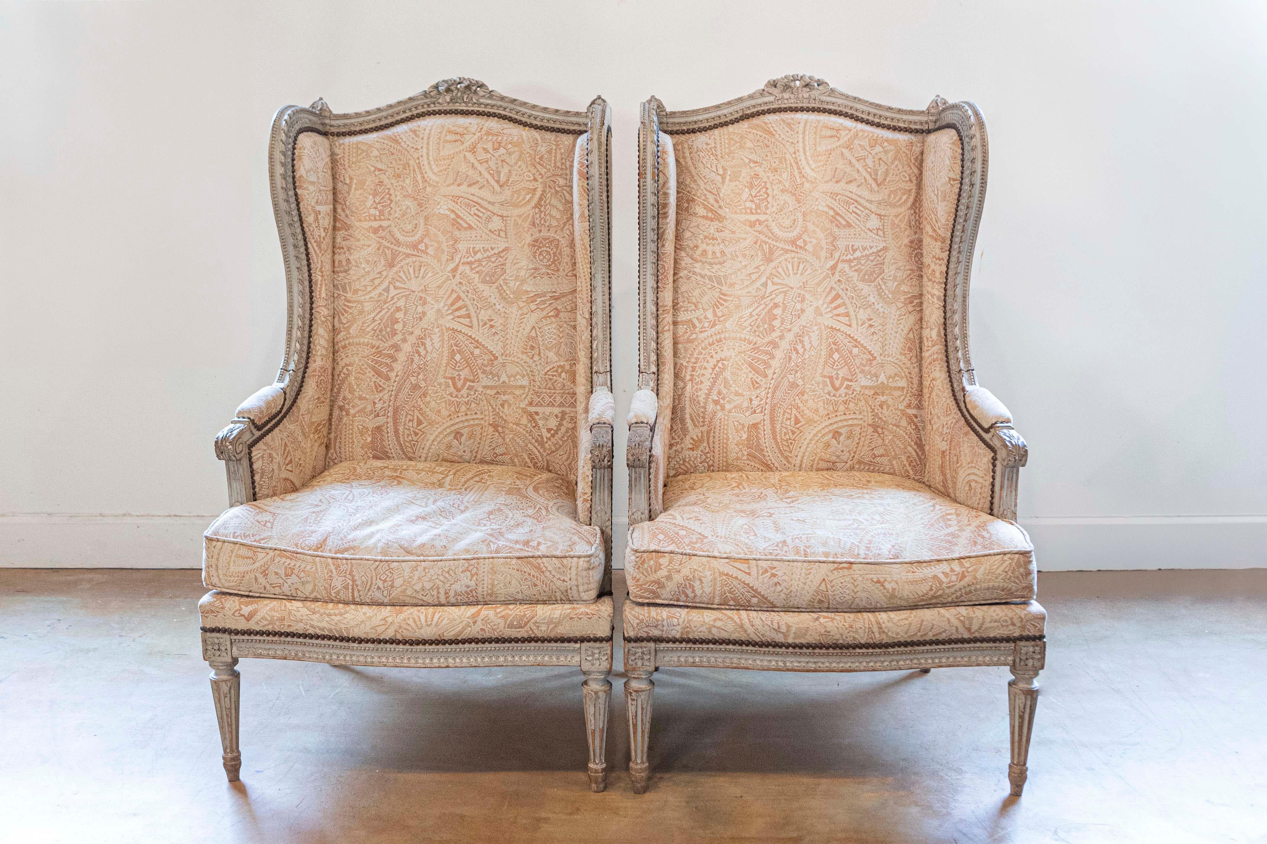 A pair of French Louis XVI style painted wood bergères à oreilles from the 19th century, with carved ribbon motifs and upholstery. Created in France during the 19th century, each of this pair of bergères features a wingback topped with a carved