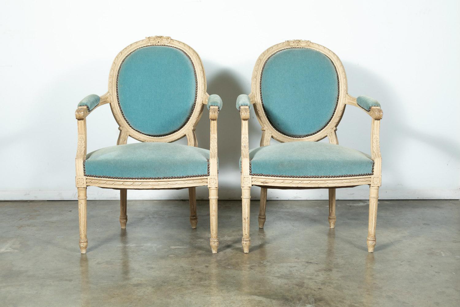 Lovely pair of Louis XVI style armchairs having medallion backs with ribbon motif and acanthus carved armrests in original cream paint with blue velvet upholstery and nailhead trim, circa 1900s. Very sturdy chairs with large seat.

Dimensions: