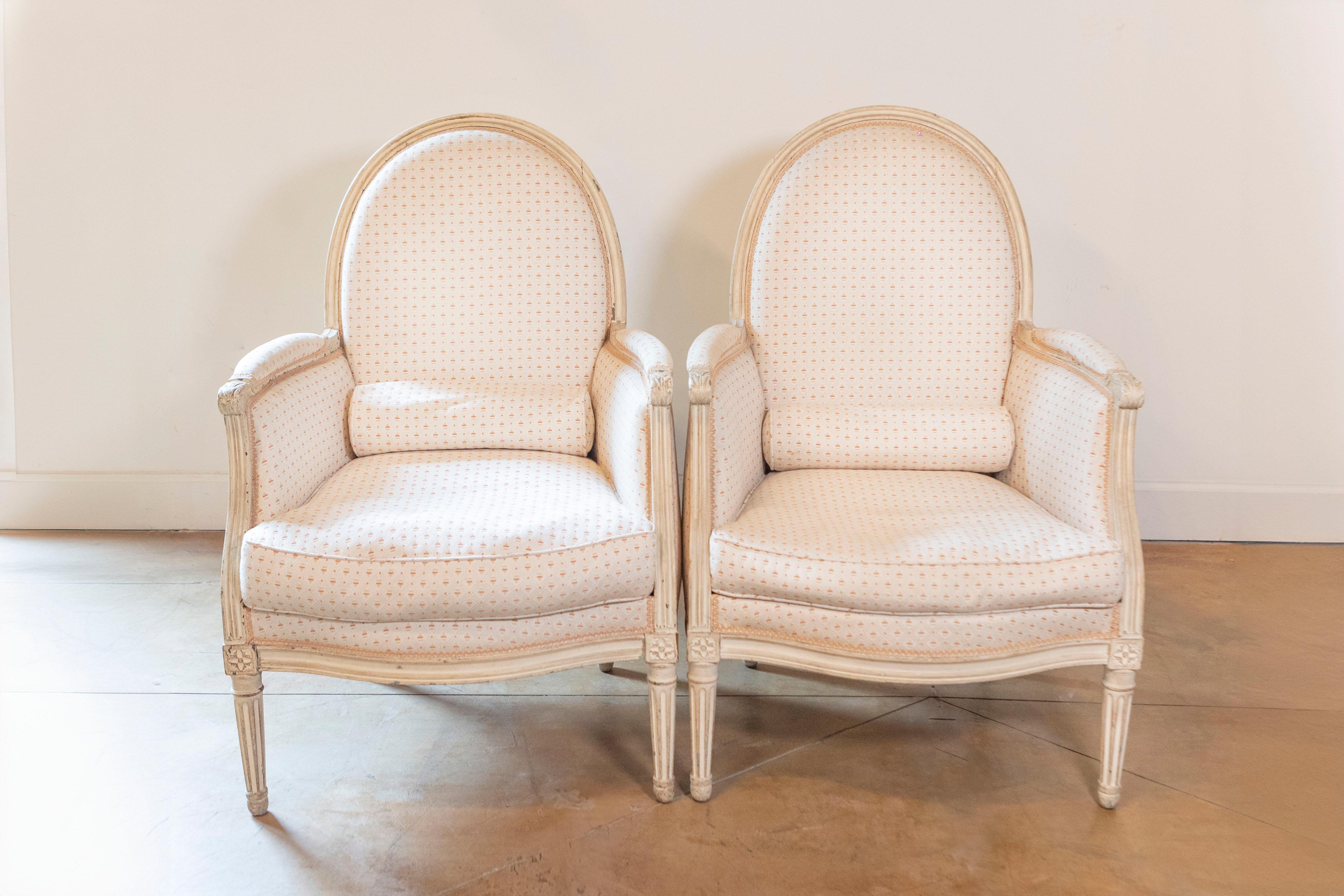 A pair of French Louis XVI style painted wood bergères chairs from the late 19th century, with oval shaped backs, scrolling arms, fluted legs and carved rosettes. Created in France during the last quarter of the 19th century, this pair of bergères