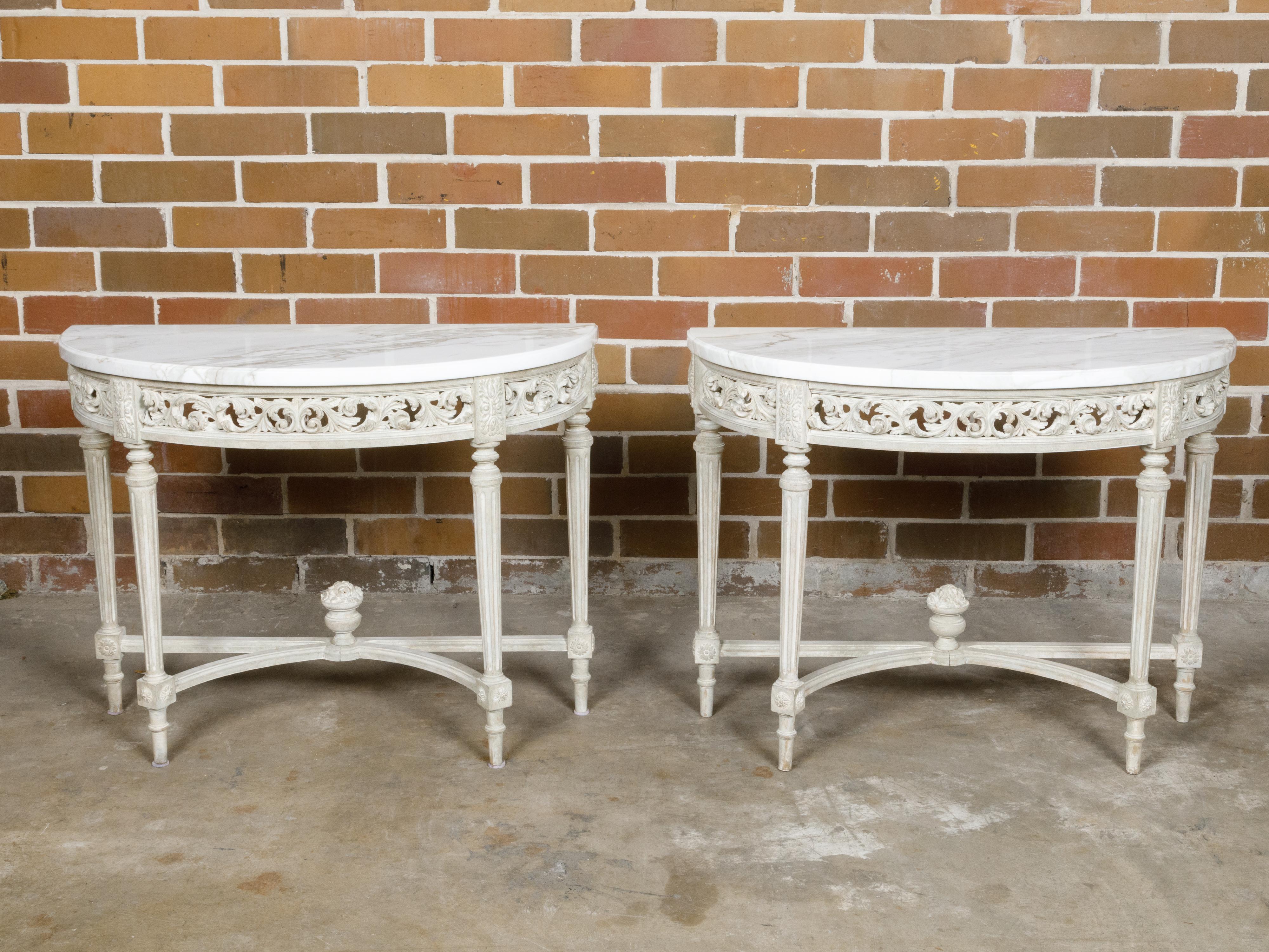 A pair of French Louis XVI style demi-lune console tables from the 19th century with white veined marble tops, carved aprons, fluted legs and arching cross-stretcher. This exquisite pair of French Louis XVI style demi-lune console tables from the