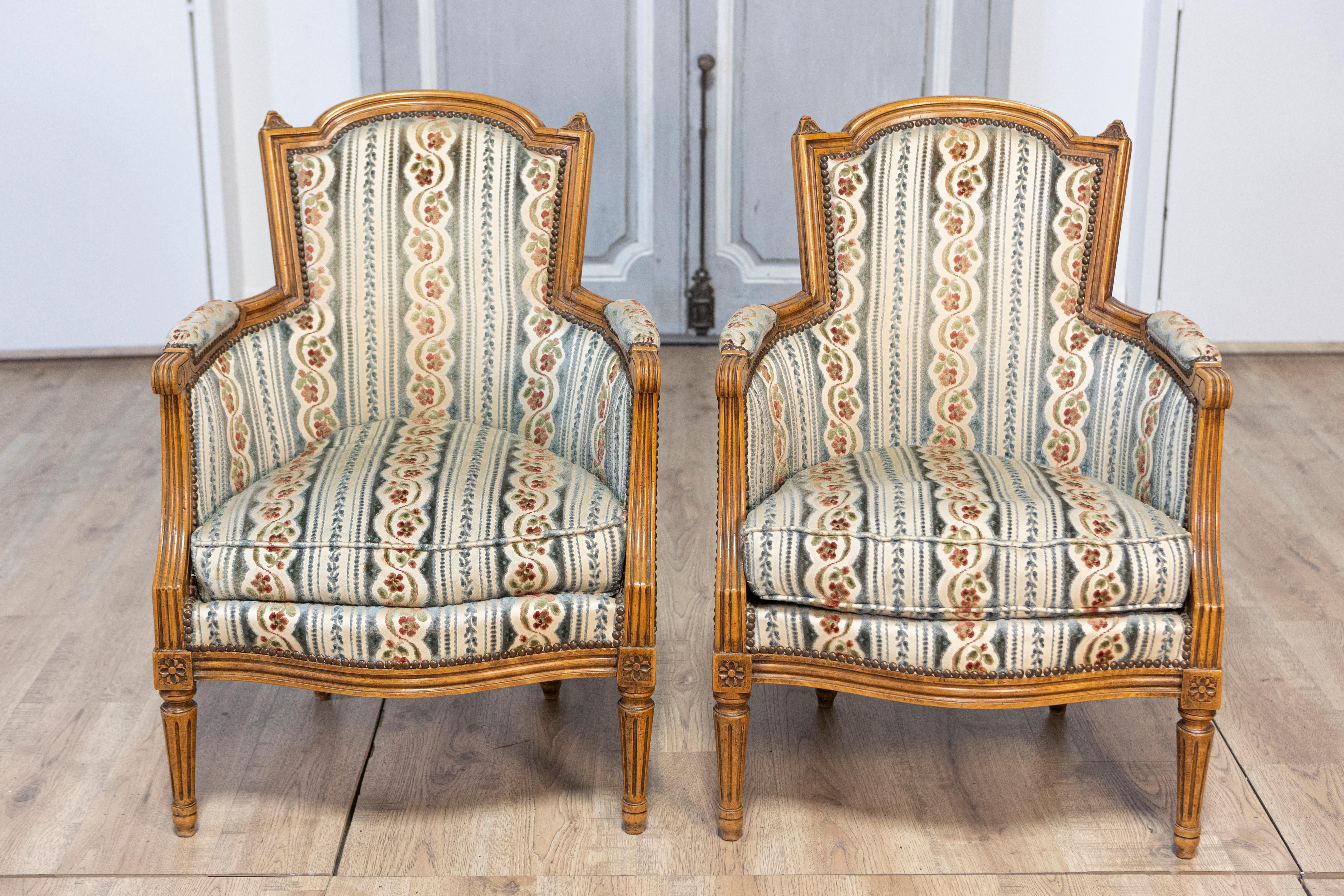 A pair of French Louis XVI style walnut bergères chairs from the 20th century with scrolling arms, carved rosettes and fluted legs. This exquisite pair of French Louis XVI style walnut bergères chairs from the 20th century embodies timeless elegance