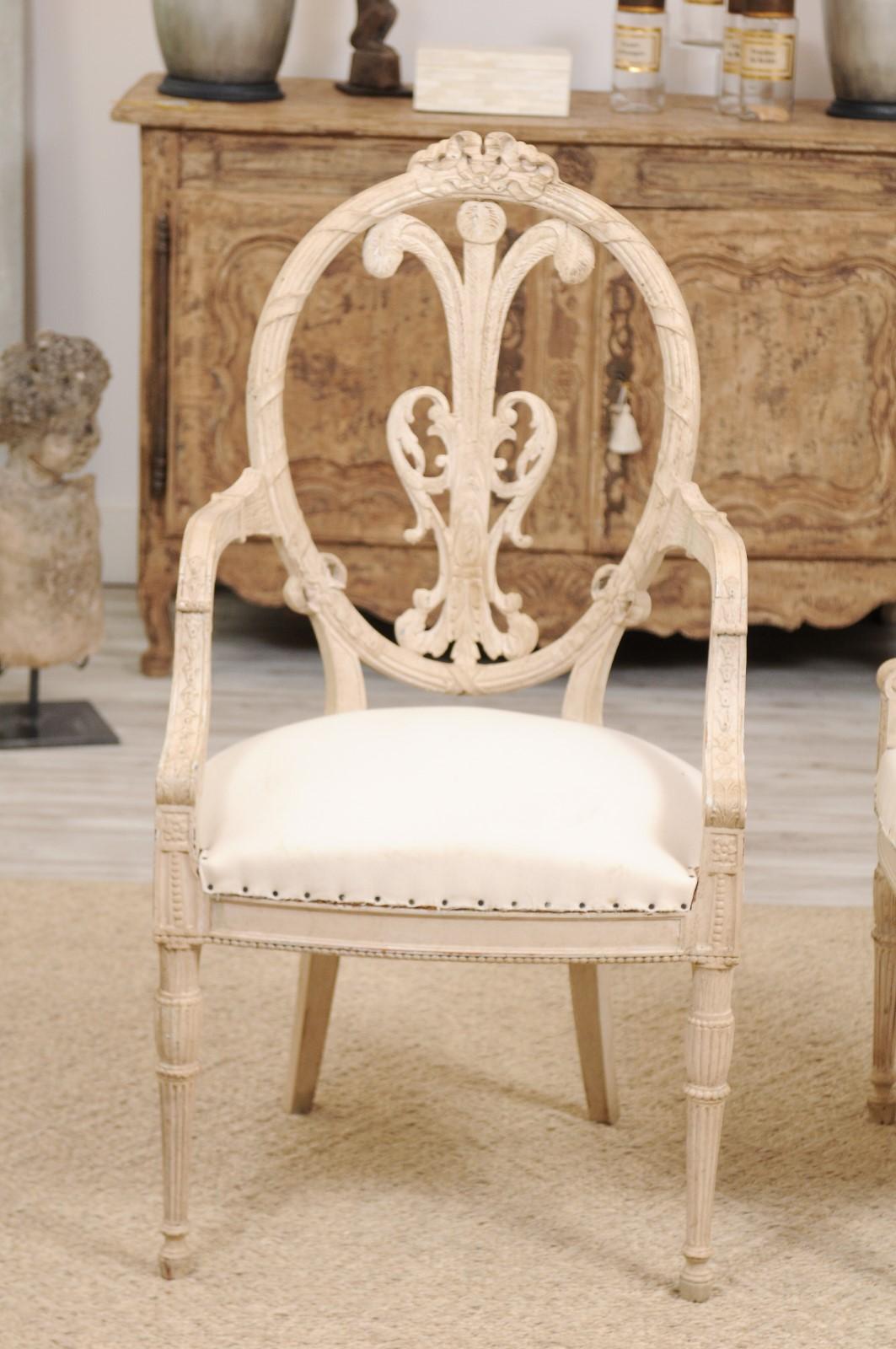 A pair of French Louis XVI style white painted armchairs from the late 19th century, with ribbon-tied oval backs, carved splats, scrolled arms, beaded motifs, fluted and saber legs. This lovely pair of painted wooden chairs feature intricately