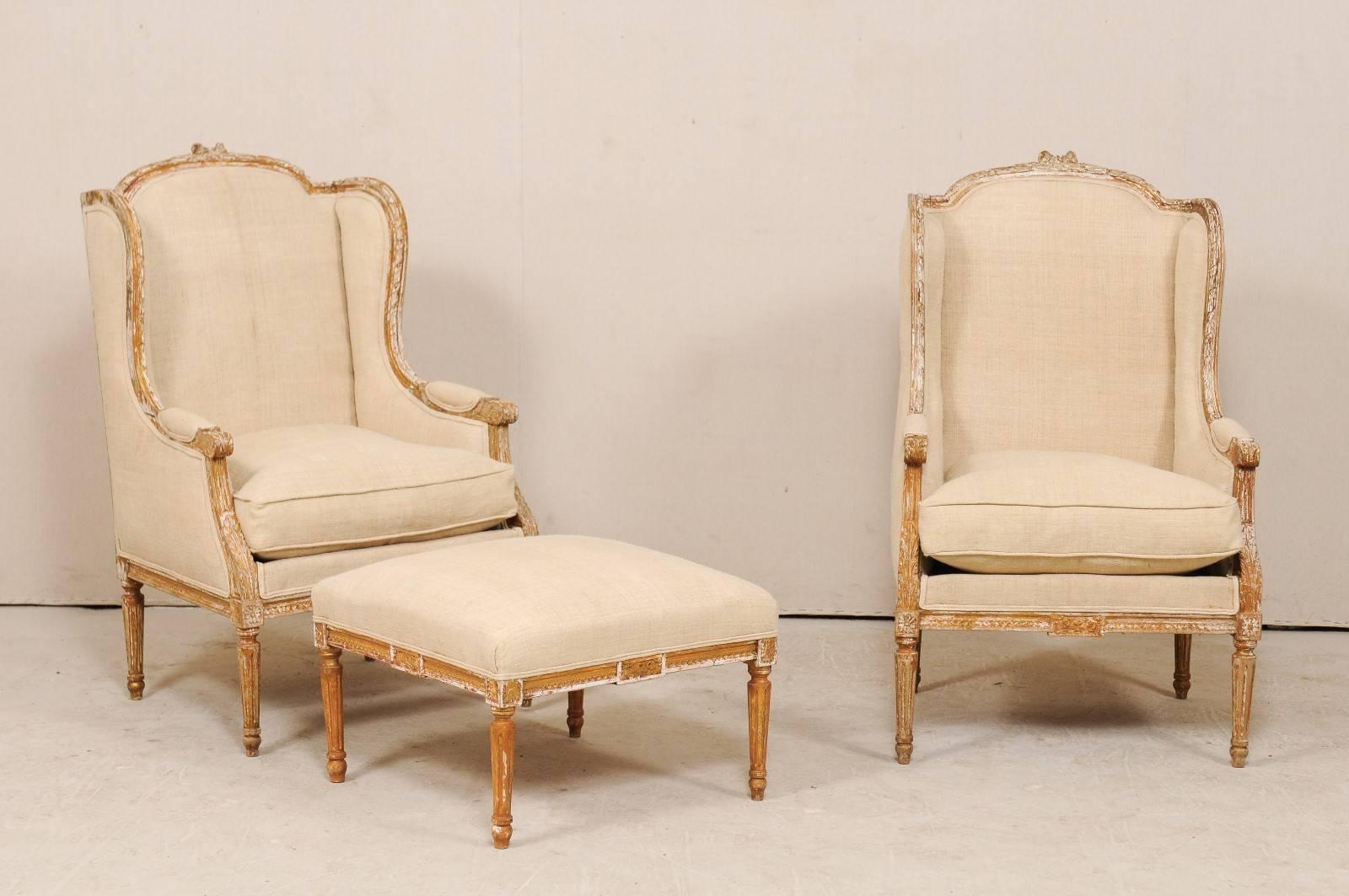 A pair of French Louis XVI style wing bergeres with single ottoman from the early 20th century. This antique French seating arrangement features two arched winged arm chairs with arched crest rails with a bow-knot at their centres, beautiful