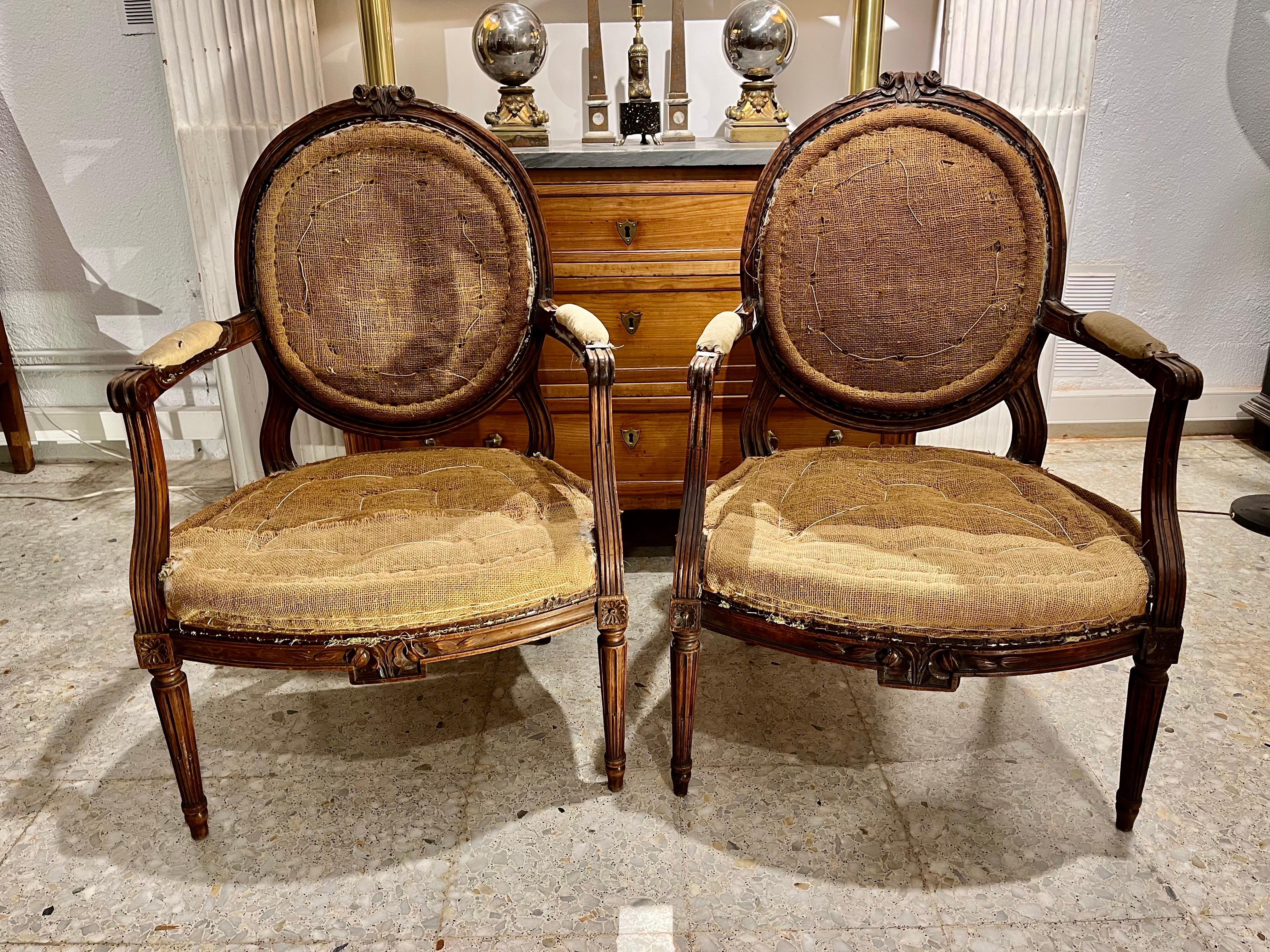 A pair of French Louis XVI-era armchairs, made of walnut wood, with hand-carved details. The chairs rest on circular fluted legs with carved rosettes at the top. The armrests are also fluted, and the oval backrest medallion ends with a floral