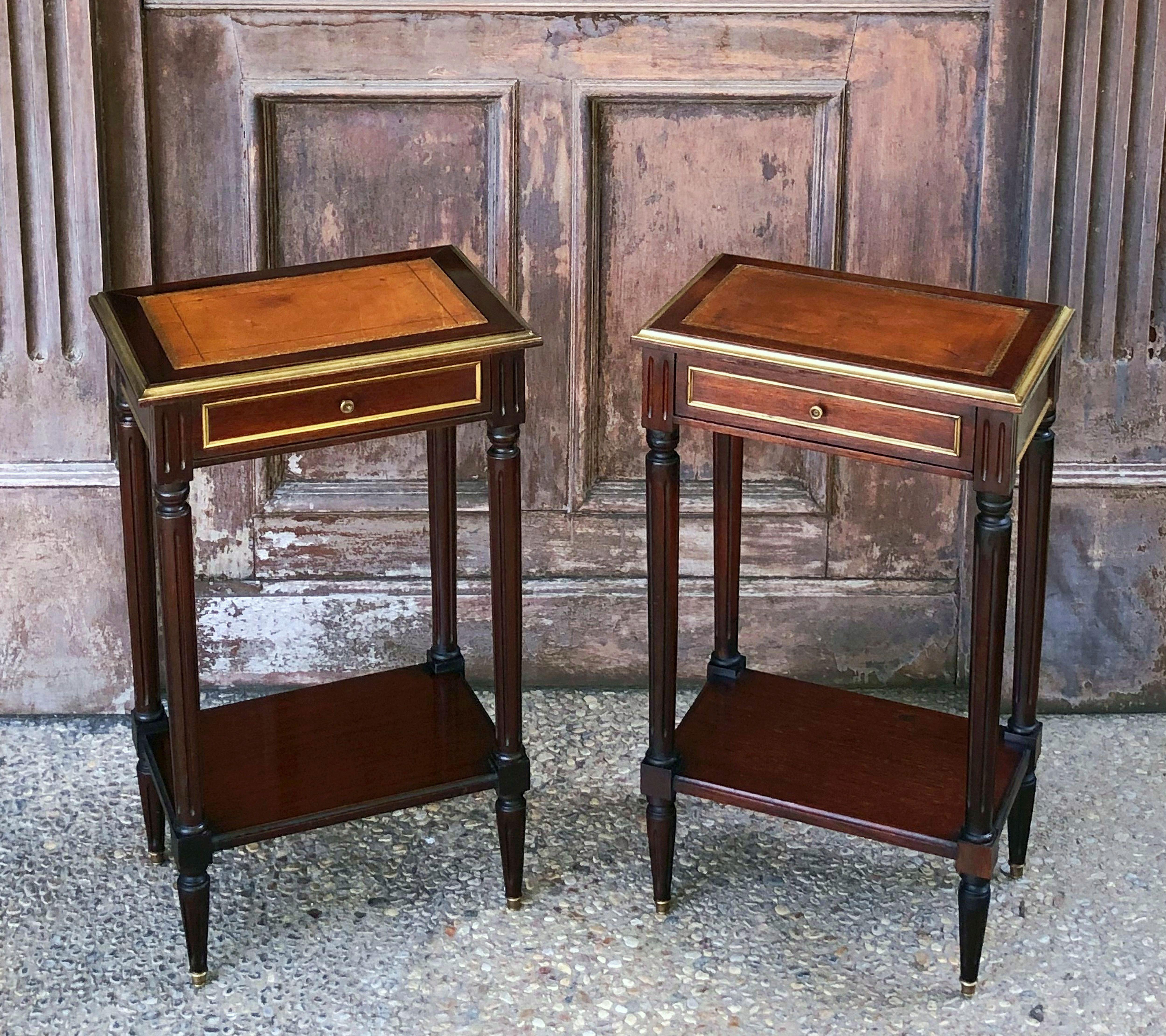 A fine pair of French side tables or nightstands (bedside tables), each table featuring a moulded rectangular top of mahogany with inset embossed leather and brass accents, over a frieze with drawer, and set upon four turned legs with lower