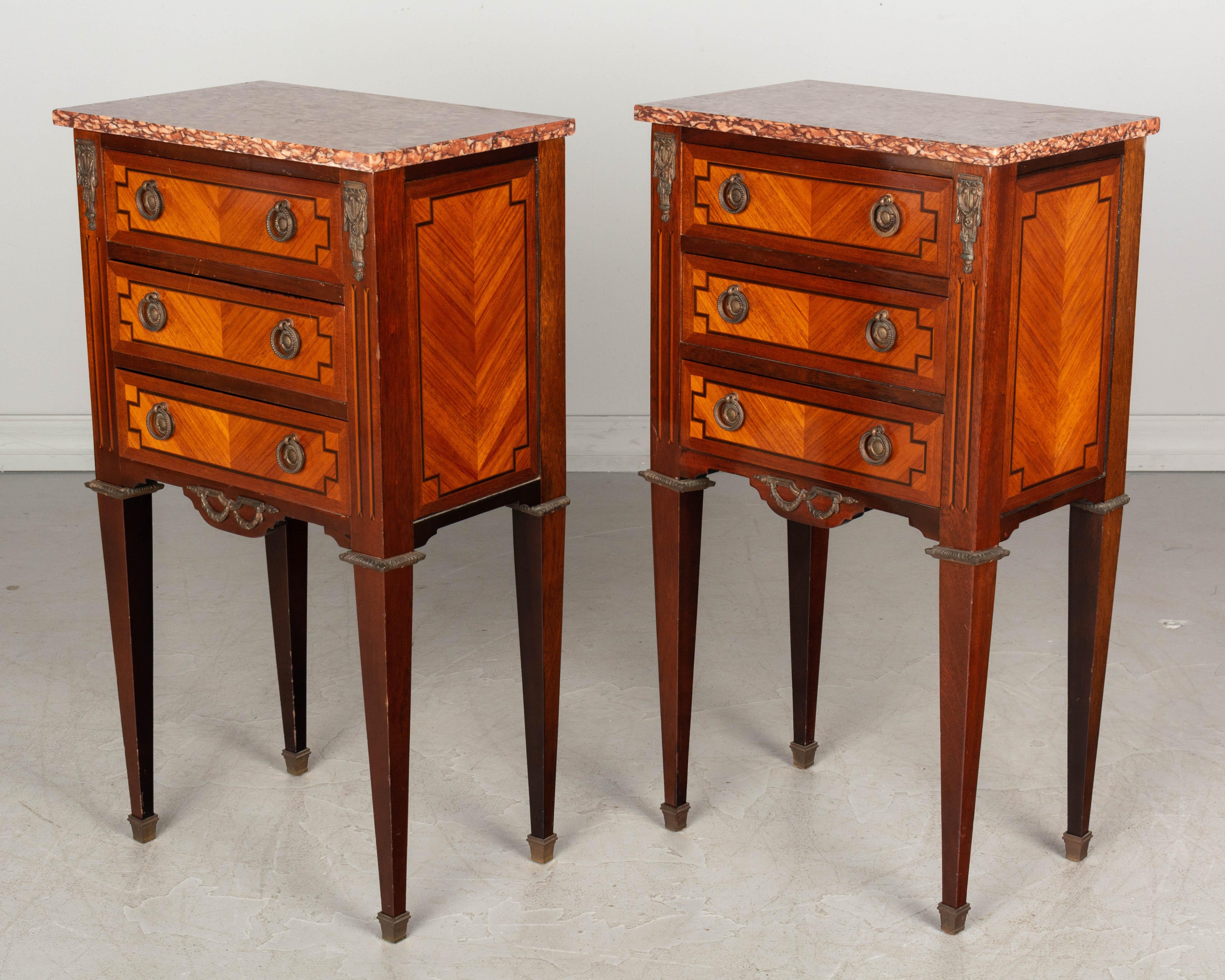 A pair of Louis XVI style French marquetry side tables, or nightstands. Inlaid veneers of mahogany and walnut with cast brass decoration. Three dovetailed drawers with ring pulls. Marble tops are not attached.