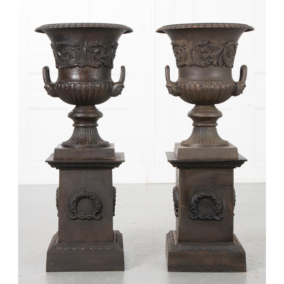A pair of urn-form planters rest upon a pair of pedestals to complete this duo of garden antiques. Made in France in the 1900’s, the planters are cast iron and each has a drain to accommodate watering. A classic shaped urn with foliage around the