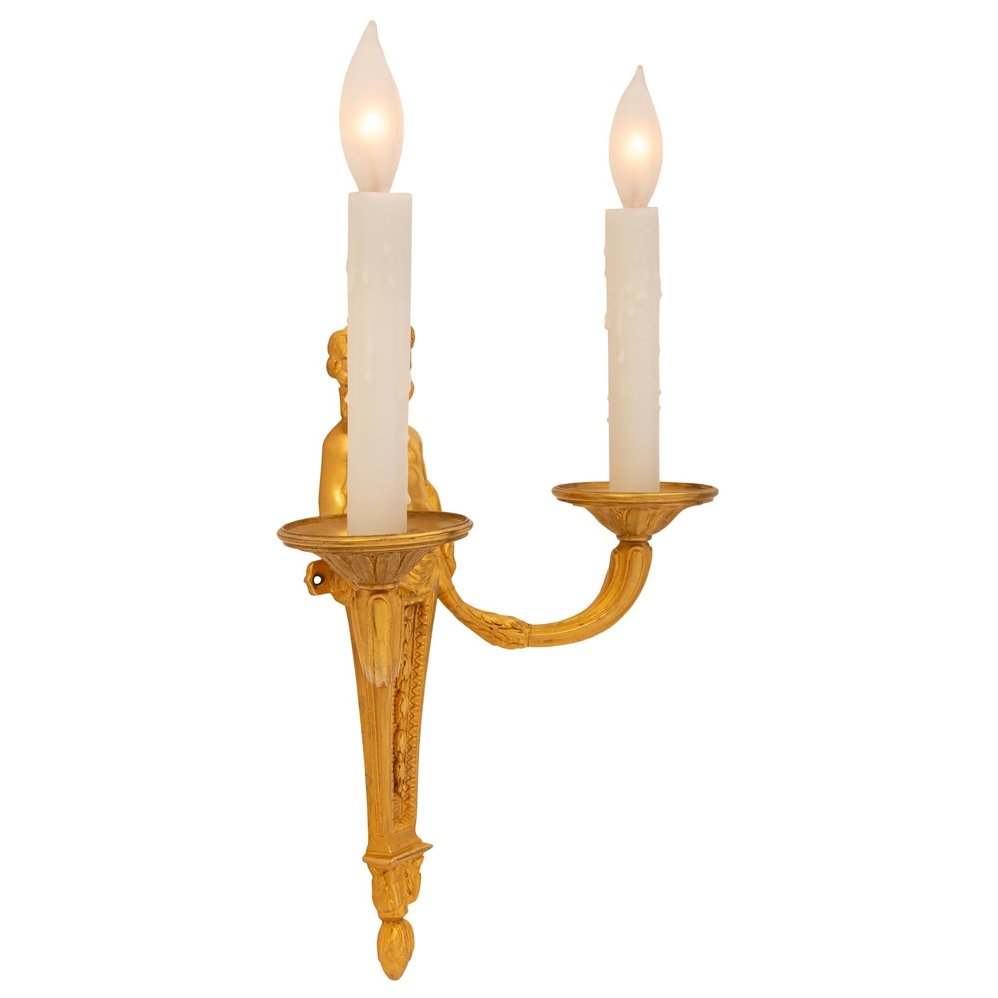Encantadora pareja de apliques franceses de ormolina de mediados del siglo XIX, de estilo Luis XVI. Cada candelabro de dos brazos está centrado por un fino remate inferior foliado bajo el elegante soporte central ahusado, decorado con ramas de