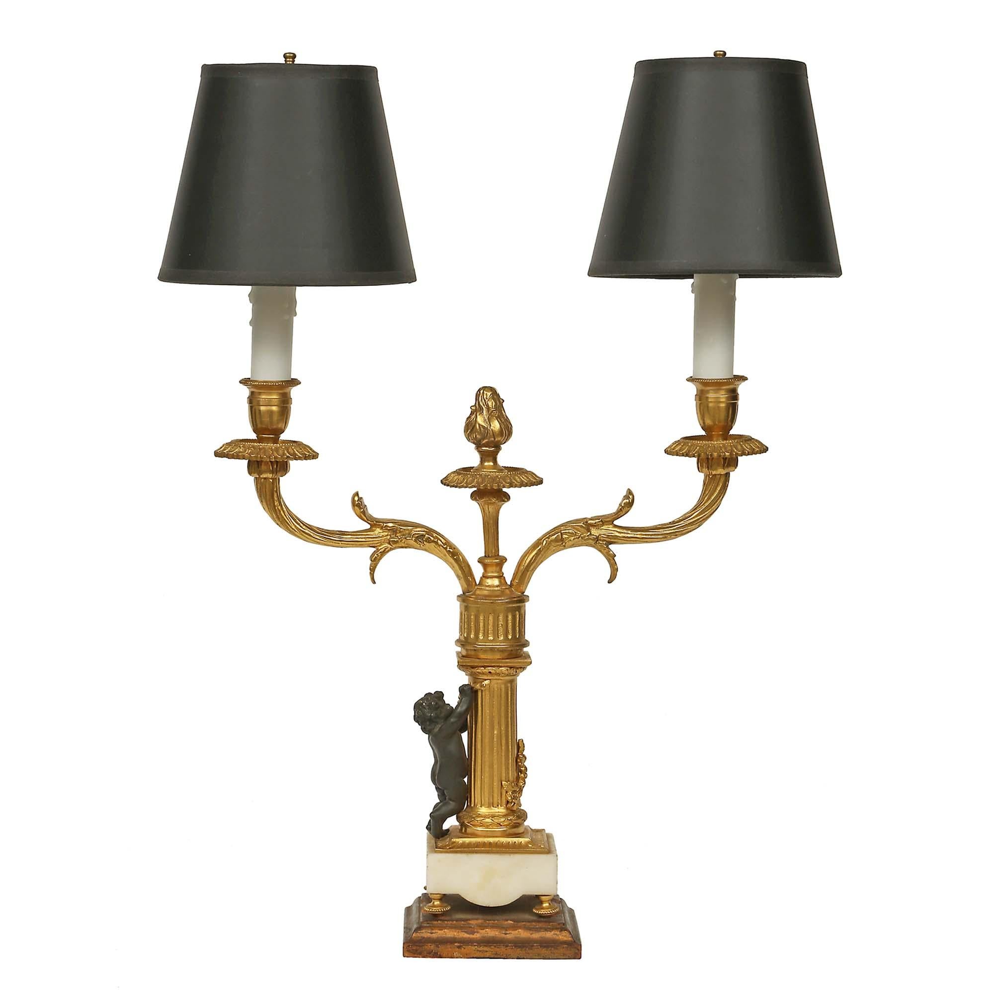 An elegant pair of French mid 19th century two arm candelabras in ormolu and patinated bronze mounted into lamps. The pair is raised on a square giltwood base below a white Carrara base. The fluted column is decorated by a bronze cherub holding a