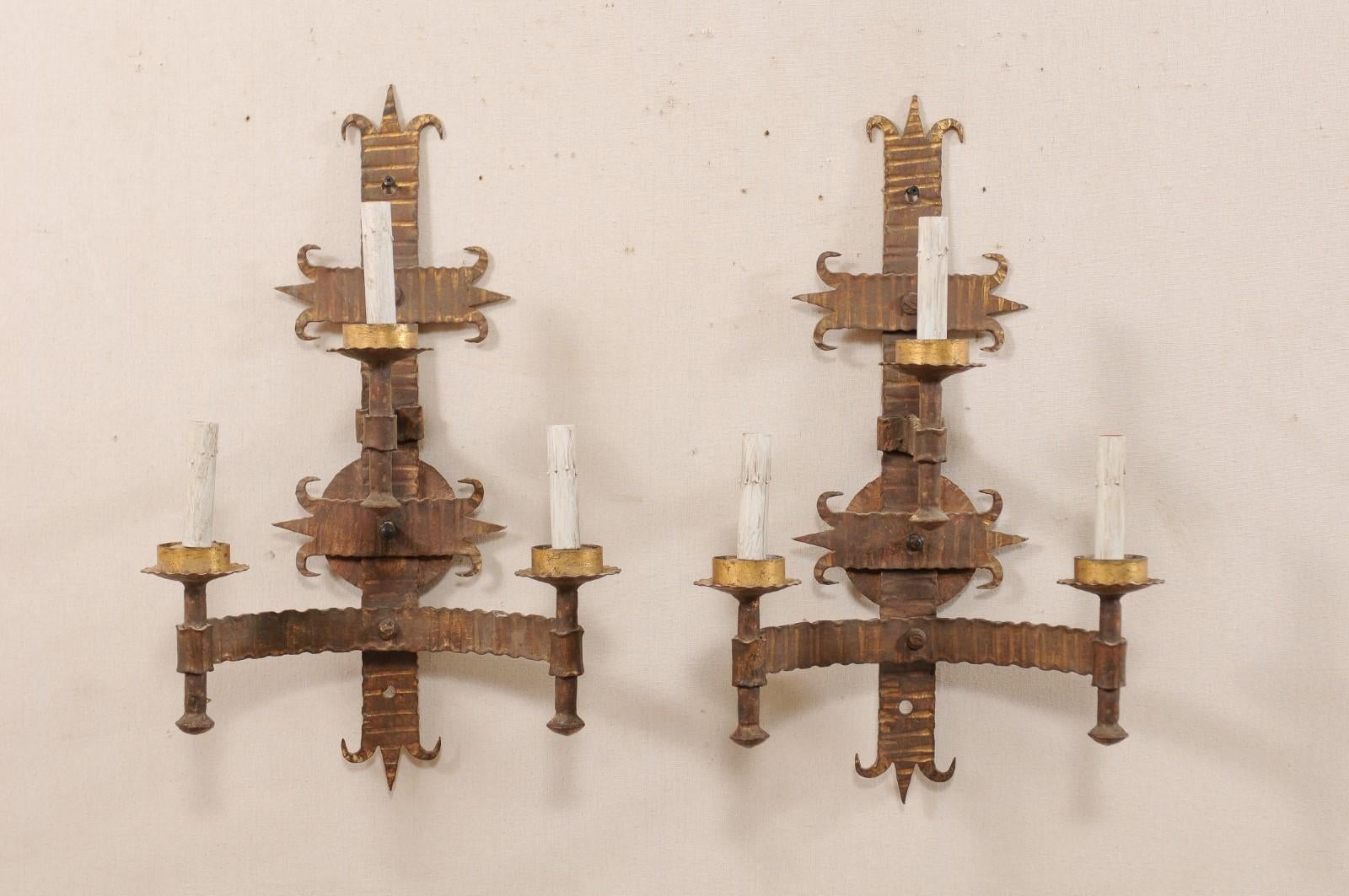 A pair of French mid-20th century forged-iron three-light sconces. These vintage French hand forged sconces each have two torch-style lights set at the farthest ends of a horizontally set C-shaped arm, with the third light, positioned higher at