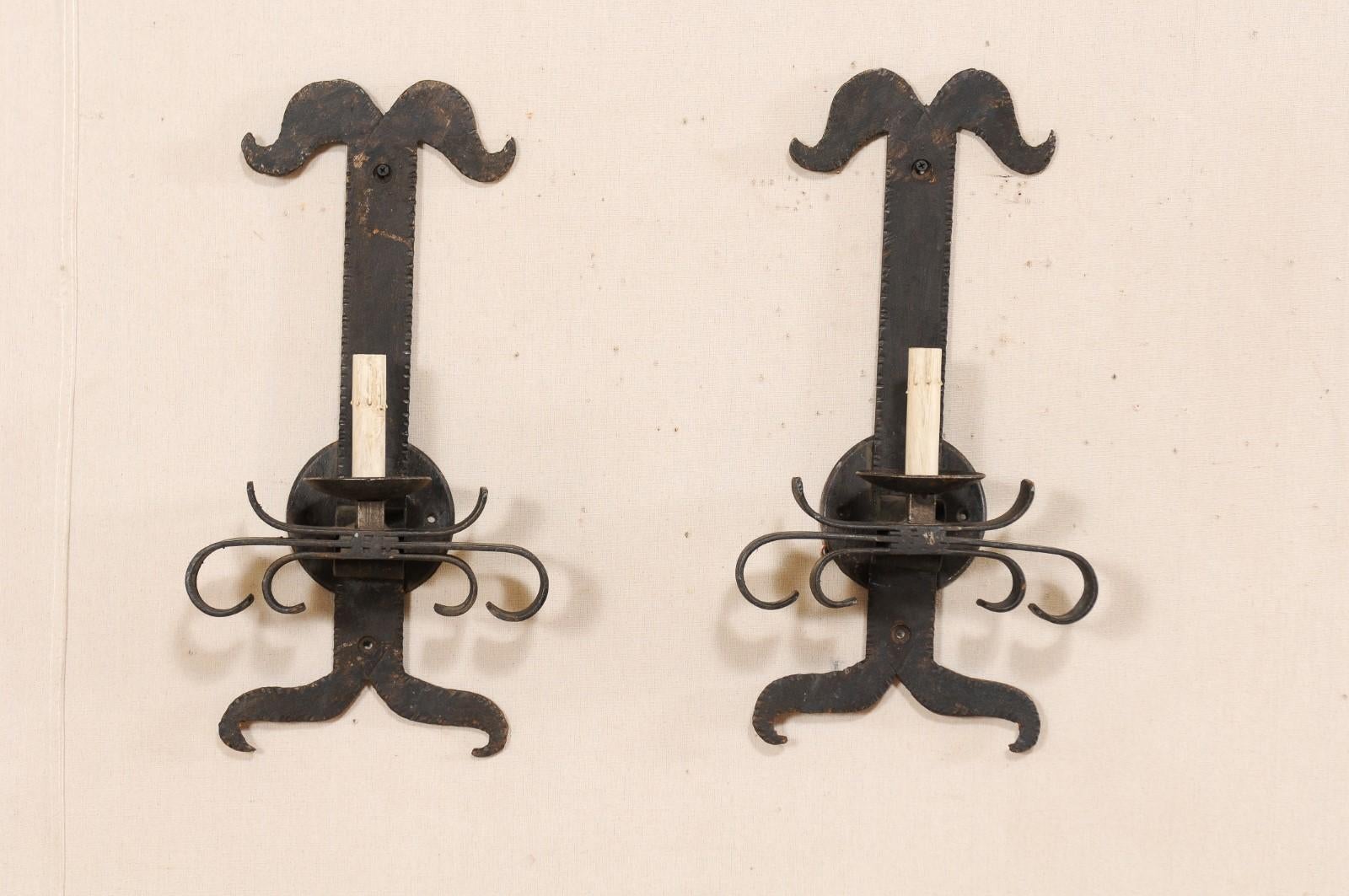 A pair of French iron single light sconces from the mid-20th century. This vintage pair of sconces from France are each made of hand forged iron and have vertically set, stylized back plates, with circular wall mount, and a single light at center