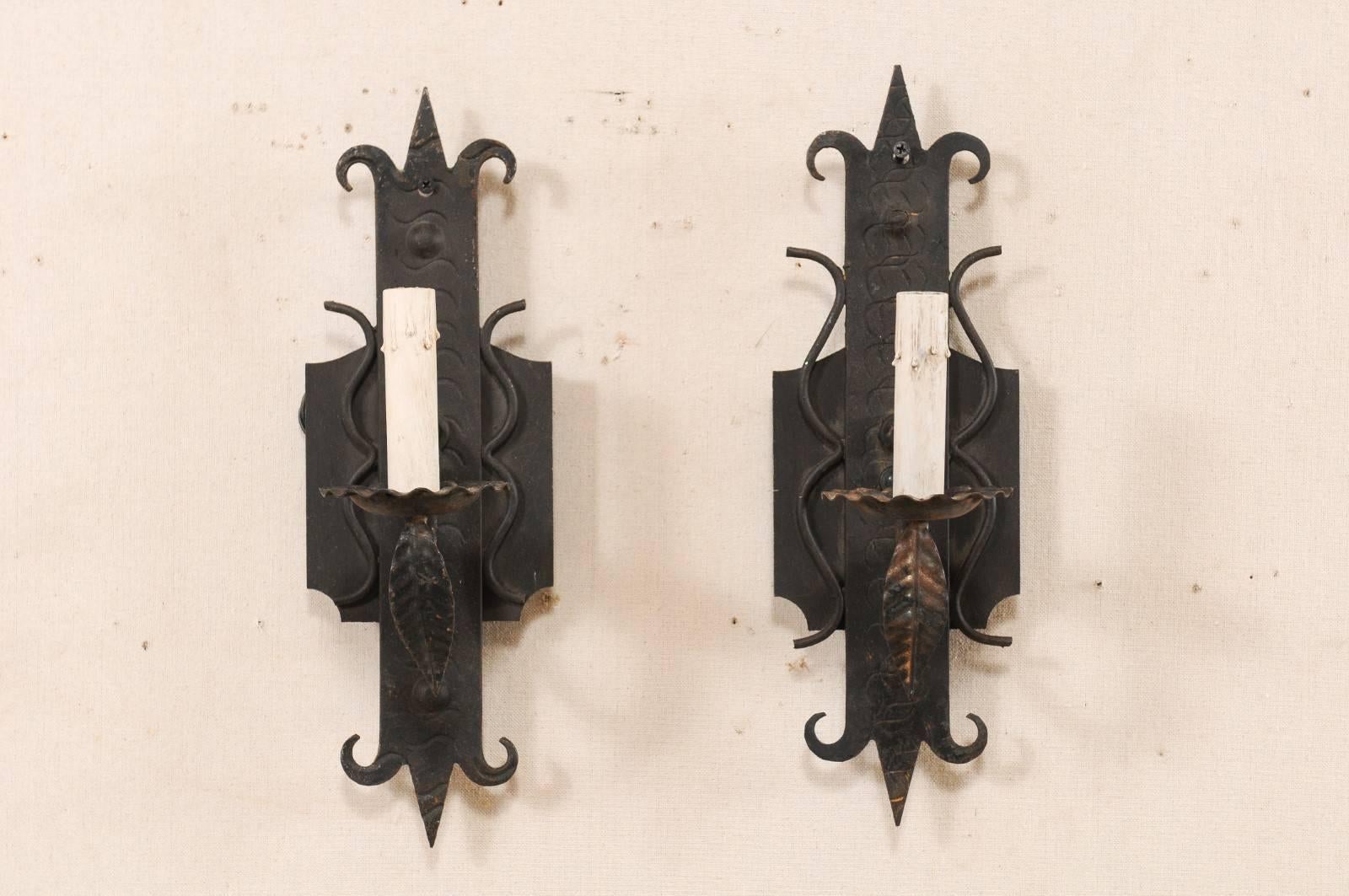 A pair of French single-light iron sconces from the mid-20th century. This vintage pair of French sconces each feature a stylized fleur-de-lys back plate with a swooped arm which extends out supporting the flower-shaped bobèche and painted candle