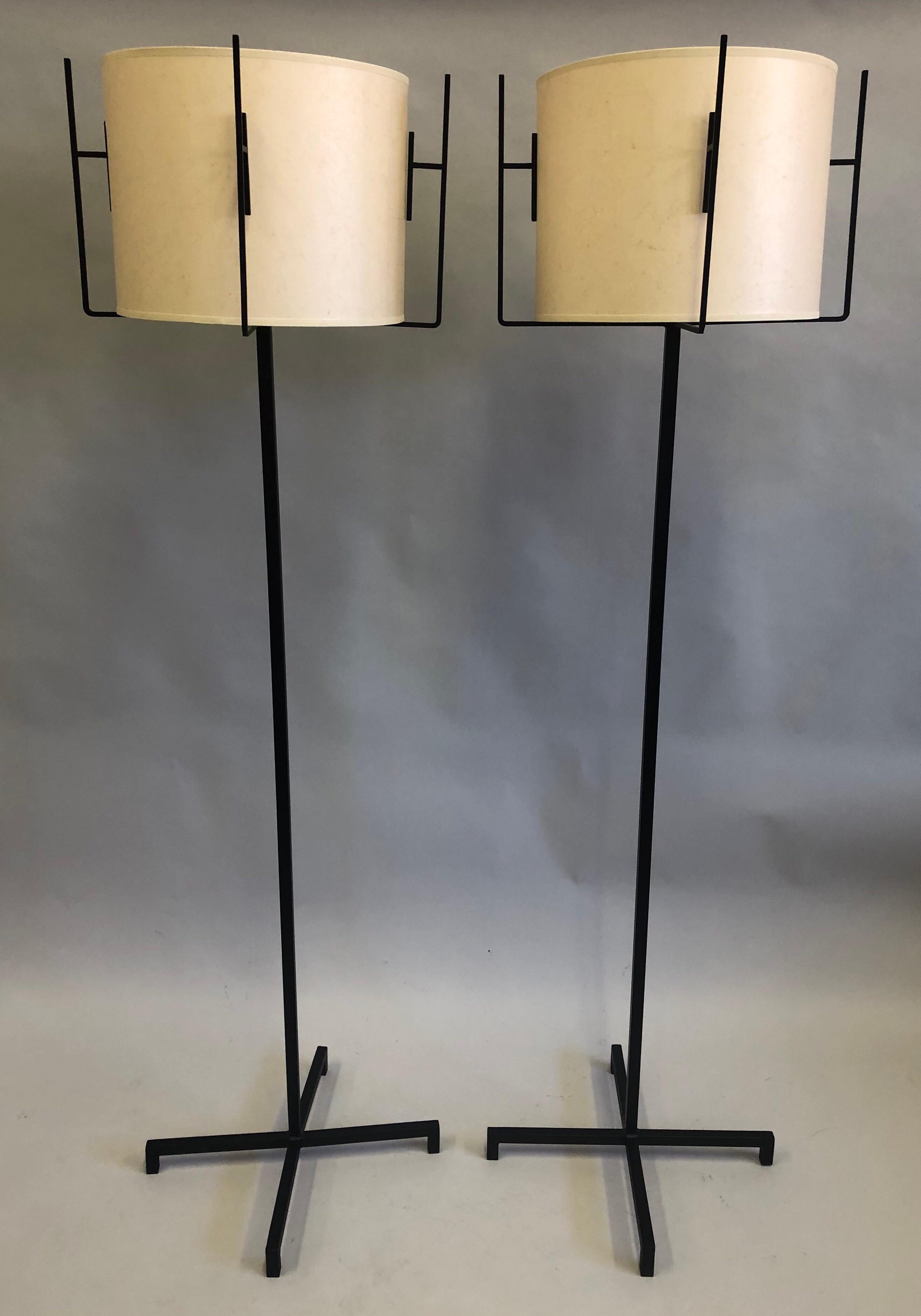 Elegant and Timeless Pair of French Mid-Century Modern Neoclassical wrought iron floor lamps by Jacques Adnet with parchment paper shades. The standing lamps have unique cross form design support system to hold the shades and this is mirrored by the