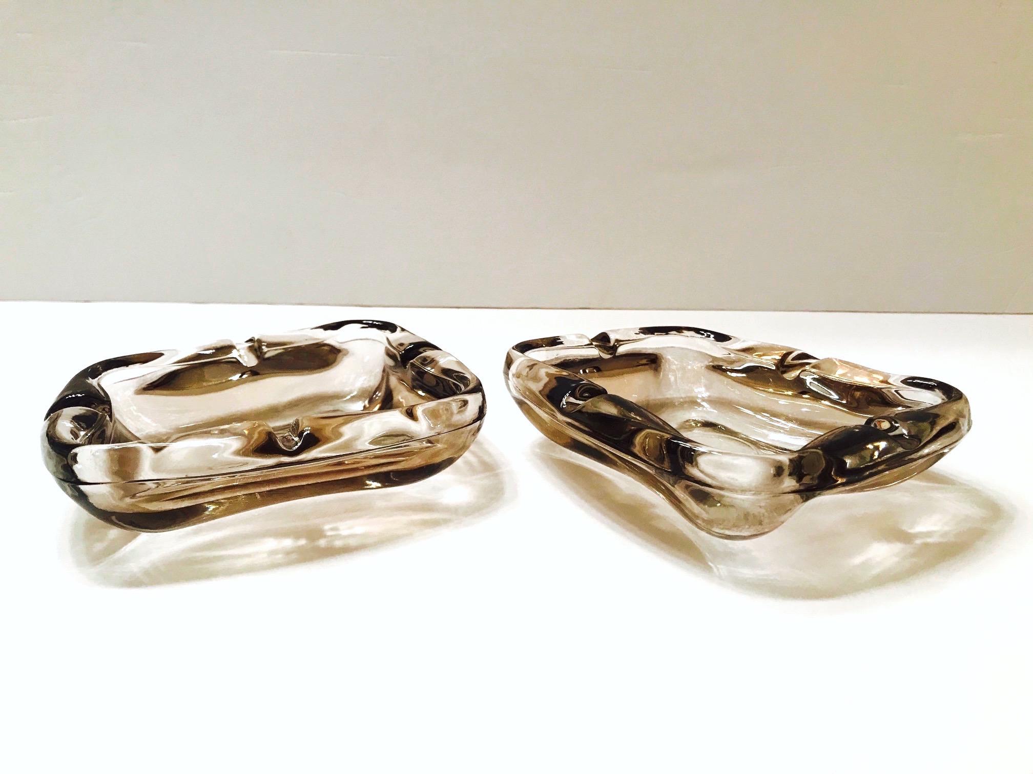 Pair of Mid-Century Modern cast glass ashtrays with handcut organic freeform design. Smoked glass in gradient hues of brown or gray depending on the surrounding light. Each ashtray is identical in scale and features four notches fitted for