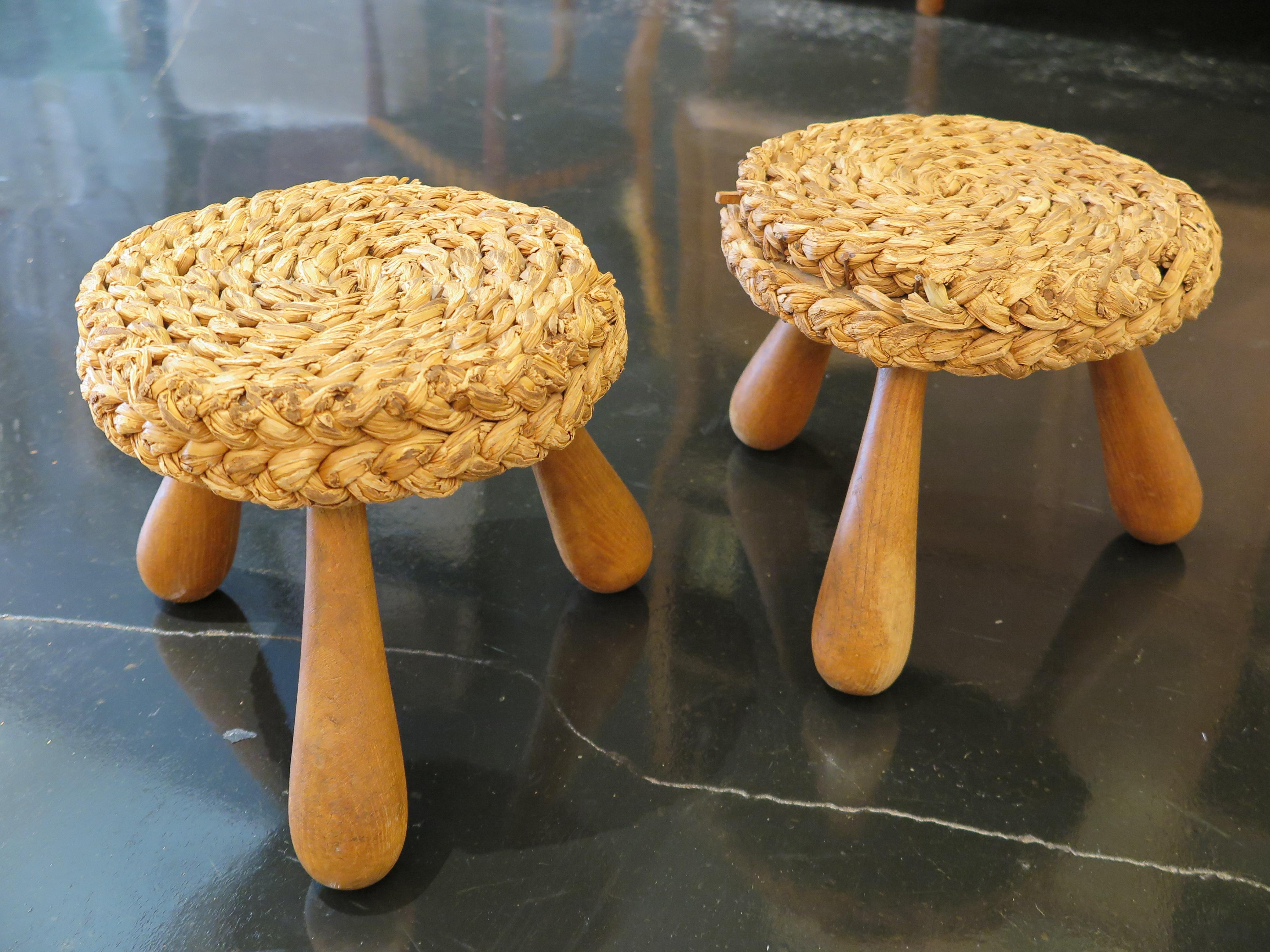 Pair of French mid-century low Shepard's stools by Adrien Audoux and Frida Minet. The rustic French poufs feature round seats in woven raffia/rattan with tripods legs and an oak keel base. Original condition.