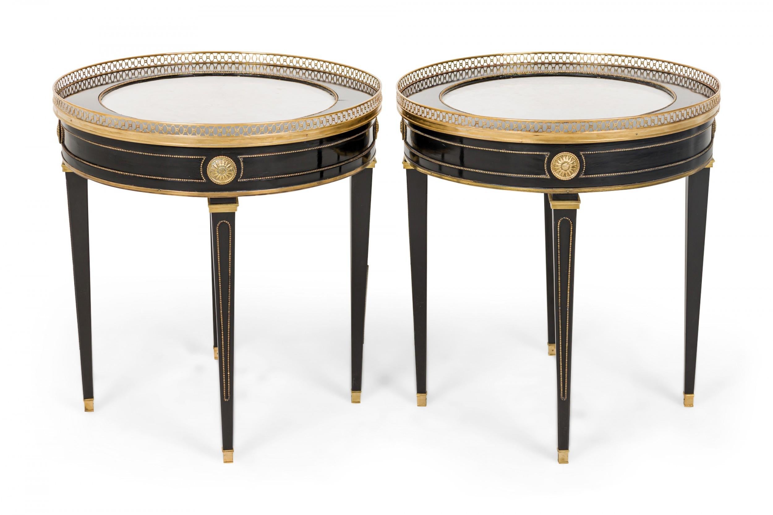 Pair of French mid-century (circa 1950) circular ebonized wood end / side tables with gilt medallions and trim and an inset white marble top surrounded by a brass gallery and resting on four tapered square legs with inset gilt detail, ending in four