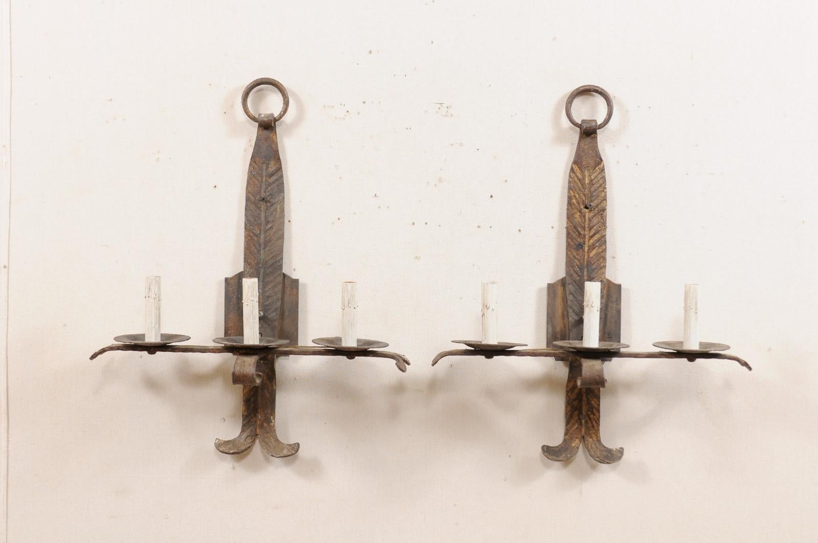 A pair of French three-light forged-iron sconces from the mid-20th century. These vintage hand forged sconces from France each feature a horizontally placed bar, with splayed tips, which provide support to the three iron bobeches with painted candle
