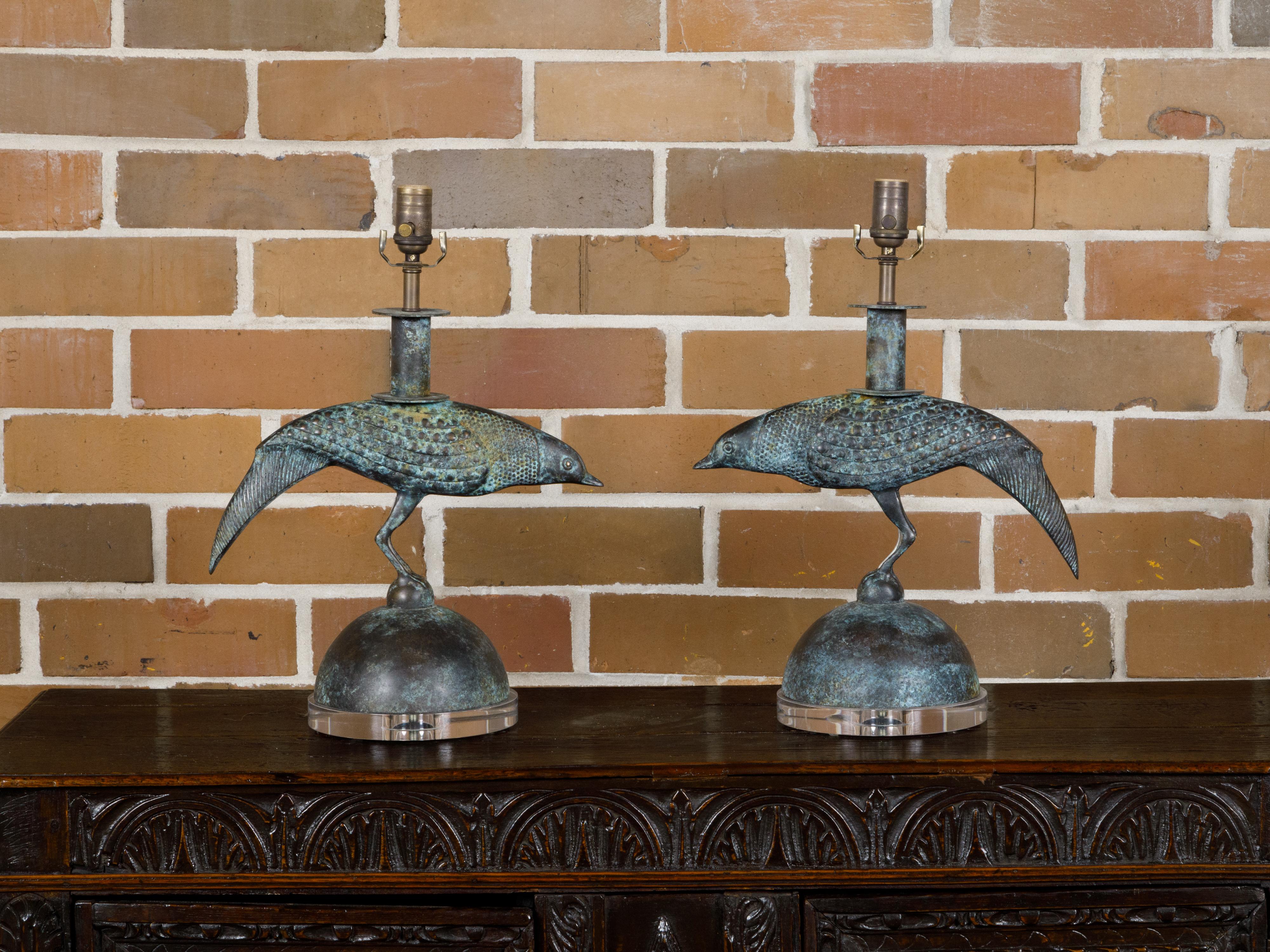 A pair of French Midcentury bronze table lamps from circa 1950 depicting birds with verdigris patina and resting on circular lucite bases. These French Midcentury table lamps, dating back to circa 1950, are a splendid representation of post-war