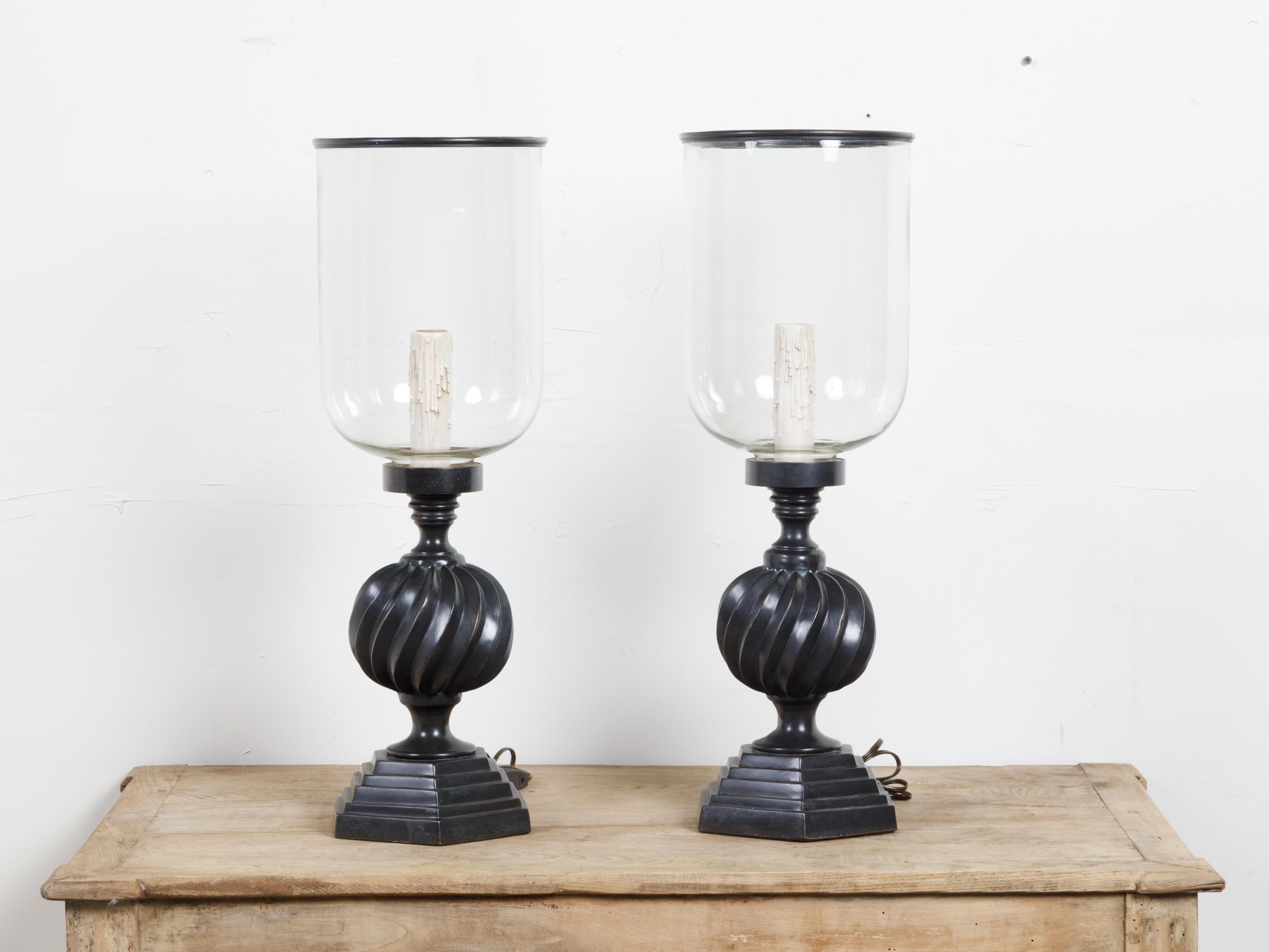 A pair of French vintage bronze hurricane table lamps from the mid 20th century, with single light, twisted motifs and stepped bases. Created in France during the midcentury period, each of this pair of hurricane lamps features a glass container
