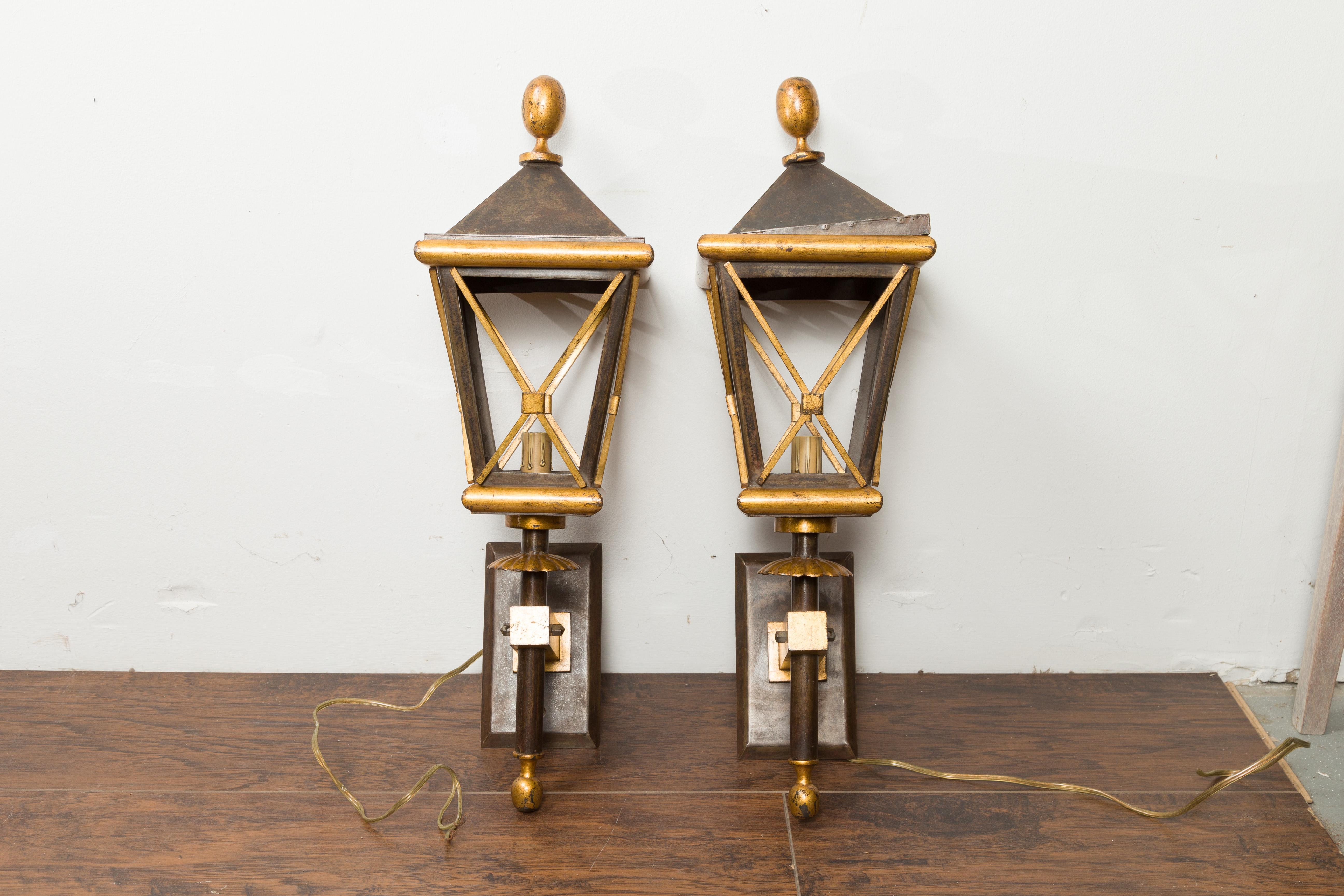 A pair of French gilt iron wall mounted lanterns from the mid 20th century, with single lights and acorn style finials. Created in France during the midcentury period, each of this pair of lanterns features a rectangular backplate connected to a