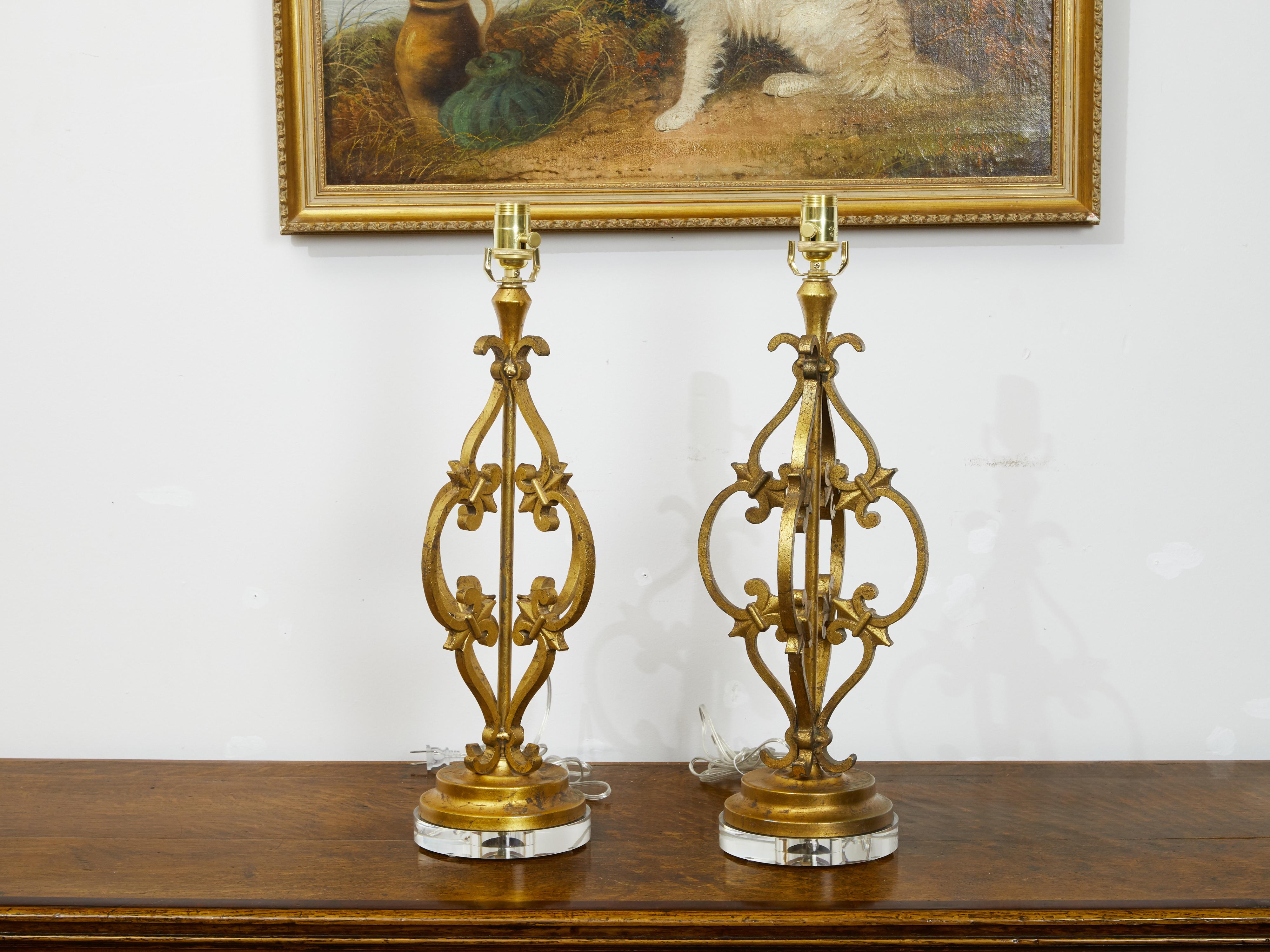A pair of French gilt metal table lamps from the mid 20th century, with scrolling effects, fleurs de lys and lucite bases. Created in France during the midcentury period, each of this pair of table lamps features a delicate arrangement of S and C